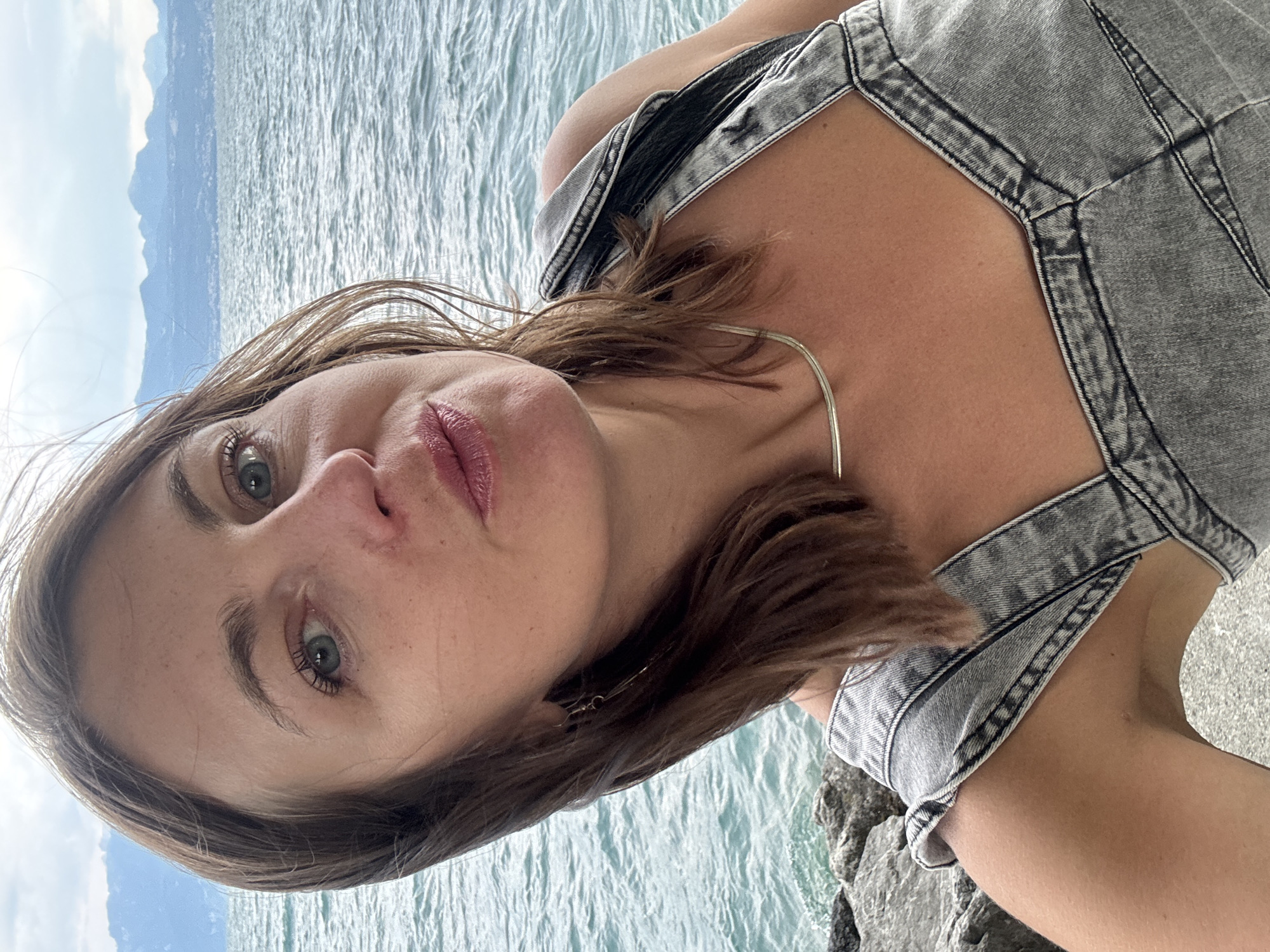 A woman with brown hair and green eyes takes a selfie outdoors, facing the camera with her mouth slightly pursed. She is wearing a grey denim top and a silver necklace. Behind her, a body of water, likely Lake Geneva, stretches out to a distant shoreline with mountains. The scene appears to be a waterfront area in Lausanne, Switzerland, with rocks visible in the foreground.