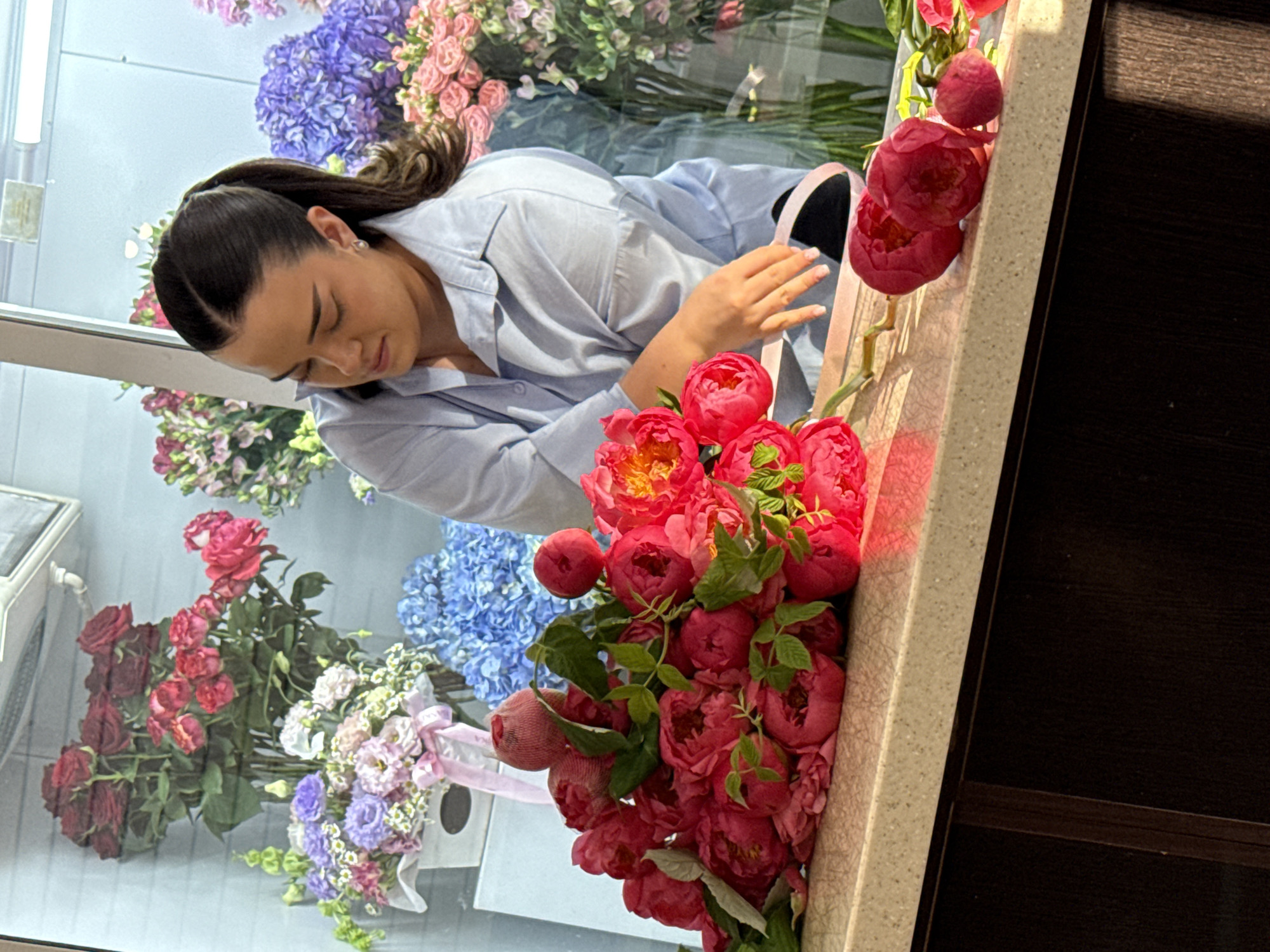 A young woman with dark hair pulled back in a ponytail, wearing a light blue collared shirt, is arranging a bouquet of vibrant pink peonies. She is leaning over a beige countertop, her hands actively working with the flowers. In the background, a display of various flowers in vases and buckets is visible through a glass window or partition. The arrangement includes red roses, purple hydrangeas, and other assorted blooms. The scene appears to be a flower shop or a similar floral arrangement setting. The specific location of Assinovskaya, Russia is not depicted in the image itself.