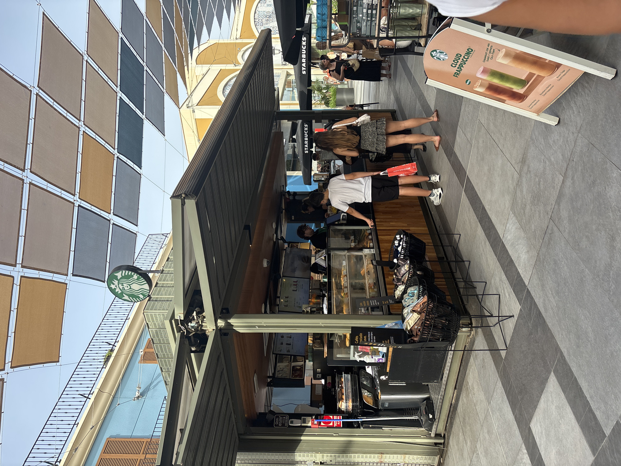 In the city of Orihuela, Spain, an outdoor Starbucks kiosk is depicted. The kiosk is under a covered patio with a multi-colored fabric canopy overhead. A Starbucks logo is visible on the top of the kiosk. A man and a woman are standing in line at the kiosk, and a woman with a black bag is walking away from it. In front of the kiosk, there are baskets of coffee beans. A sign in front of the kiosk advertises "Cloud Frappuccino" with images of three different colored drinks. The ground is paved with large, grey tiles in a geometric pattern. The background shows buildings with arched windows and a blue wall with brown shutters. The sky is visible through the canopy.