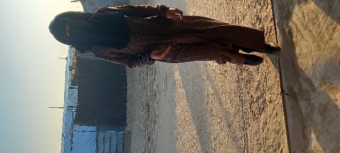 A person clad in a long brown coat and wearing a black niqab stands on a sandy surface. Behind the individual are rows of tents or temporary shelters. The ground shows textured patterns possibly from tire tracks or sand drifts. The lighting appears to be from a low sun angle, creating long shadows. The overall impression suggests an encampment in a dry, open area.