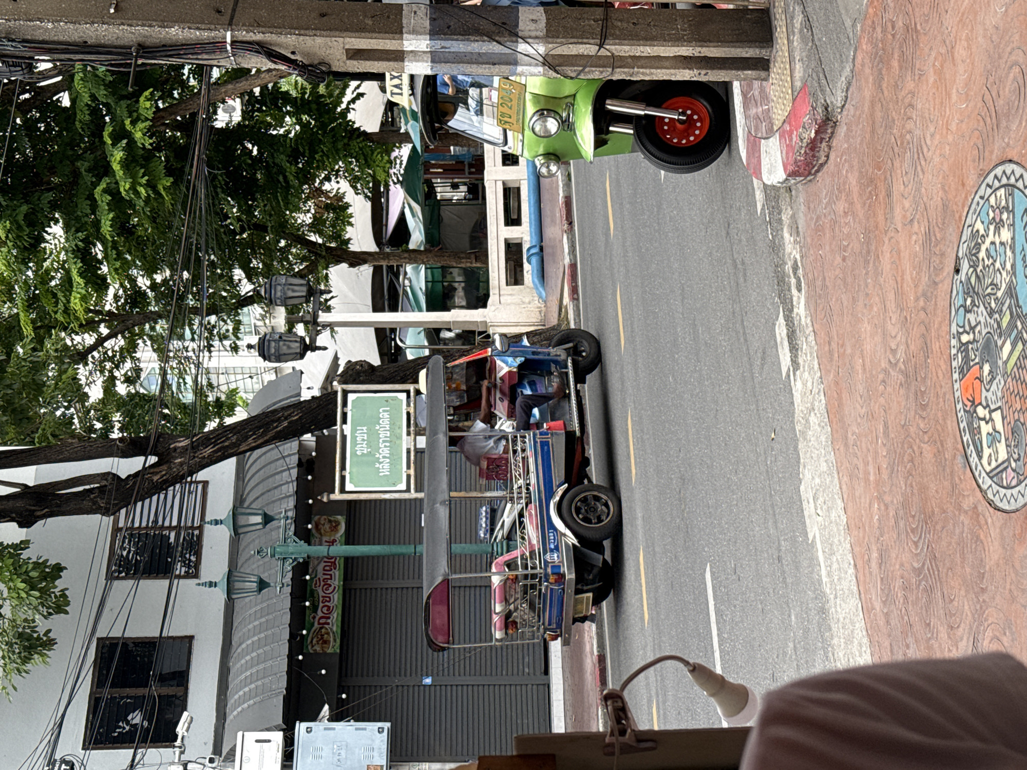 In Bangkok, Thailand, a street scene unfolds with a blue and red tuk-tuk in the foreground, its driver partially visible. To its right, a green tuk-tuk, labeled "TAX" on its roof, is parked, with its license plate reading "99 2049". The tuk-tuks are on a paved street marked with yellow lines. Buildings with gray shutters line the street, and a lamppost with two lights stands to the left. Overhead, power lines crisscross between buildings and trees. The ground in the foreground features a decorative sidewalk and a manhole cover with colorful illustrations. The overall atmosphere is that of a bustling urban environment.