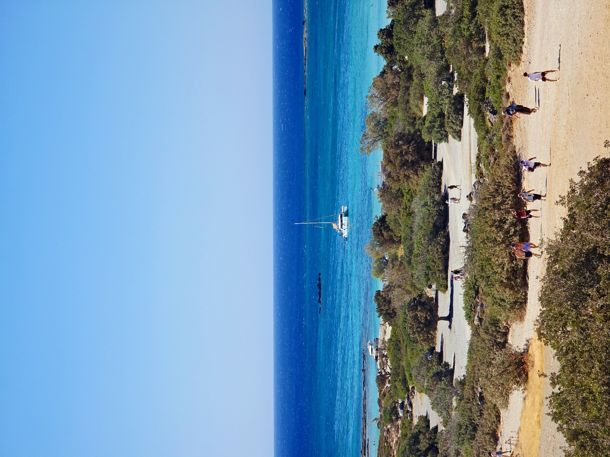 The image depicts a wide, panoramic view of a coastal landscape under a clear blue sky. In the foreground, a sandy path winds through dense, low-lying Mediterranean vegetation, interspersed with some taller trees. Several individuals, casually dressed in summer attire, are walking along the path. Some are walking individually, while others are in small groups, suggesting a relaxed, leisurely atmosphere.
Further into the middle ground, the vegetation gives way to a shallow, turquoise sea, which transitions to a deeper, vibrant blue further out. The water appears calm and inviting. A white catamaran with a single mast is anchored or sailing in the bay, its sails furled. In the distance, a rocky outcrop emerges from the sea, and the horizon is marked by a faint outline of land. Small boats are also visible near the shore, indicating a popular tourist or recreational area. The overall scene conveys a sense of a sunny, peaceful day at a coastal resort or beach. The specific location in Greece is not provided, but the vegetation and sea color are characteristic of the Mediterranean region.