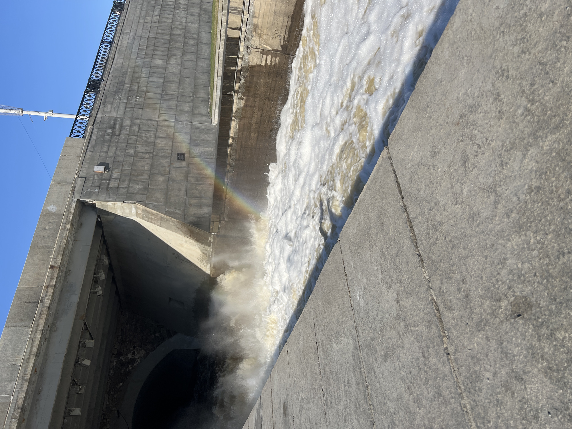 The image captures a powerful display of water management infrastructure in Yekaterinburg, Russia, under a clear blue sky. The central feature is a massive concrete dam or spillway, from which a large volume of water cascades downwards. This water is highly turbulent and frothing, appearing white and light brown due to aeration and possibly sediment.
A striking detail is a vibrant, multi-colored rainbow arcing through the mist generated by the falling water, situated above the main surge.
The concrete structure is predominantly gray, featuring large, rough-textured blocks and smooth surfaces. On the left side of the frame, a dark, cavernous arched opening is visible, possibly an outlet or tunnel, with multiple floodlights mounted within its upper interior. Above this section, a robust concrete wall is topped with a decorative black metal railing featuring a repeating circular motif. A white utility pole or mast is also visible against the clear sky in the upper left.
The foreground on the right consists of a flat, light gray concrete surface, likely a walkway or viewing platform, showcasing its textured finish and a distinct seam. The bright sunlight and clear conditions suggest it is daytime. No people are visible in the scene, and no legible text can be identified on any of the structures. The overall impression is one of immense engineering and natural beauty coexisting.
