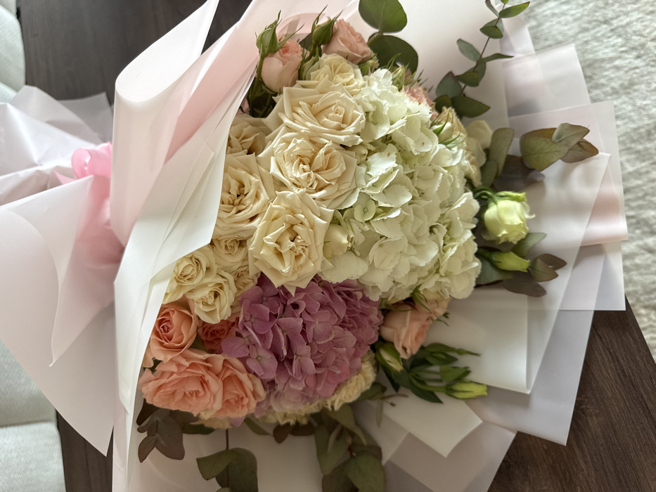 The image displays a large, professionally arranged floral bouquet, presented at an oblique angle from above. The bouquet is predominantly composed of various cut flowers, including numerous light cream and peach-colored roses, one large cluster of light purple hydrangea, and another large cluster of white hydrangea. Interspersed within the arrangement are several sprigs of eucalyptus leaves, characterized by their rounded, grey-green foliage. The flowers are wrapped in multiple layers of translucent white and light pink paper or cellophane, secured at the top by a light pink ribbon. The lower portion of the bouquet rests on a surface partially covered by a light-colored, possibly textured fabric or rug, with a darker, wood-grain-like surface visible on the left side of the frame. No individuals are present. The image contains no discernible elements to determine its precise location within the city of Dubai, United Arab Emirates.