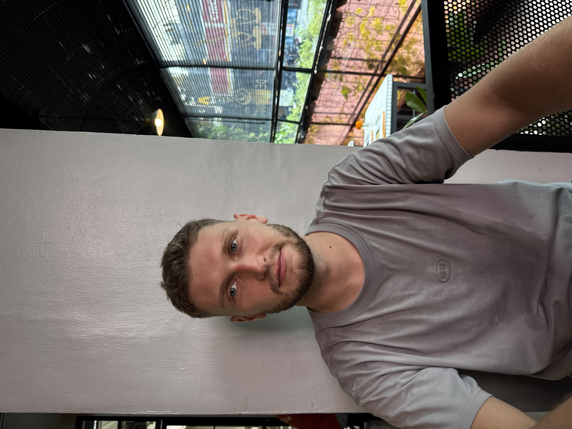 A man with short brown hair and a beard is wearing a grey t-shirt. He is indoors with a view of the outside through a ceiling composed of a grid of glass or transparent panels, which reflect the cityscape. The environment appears to be a café or restaurant in an urban area, possibly North Kuta, Indonesia. A white wall is visible on the left. The man is looking directly at the camera.