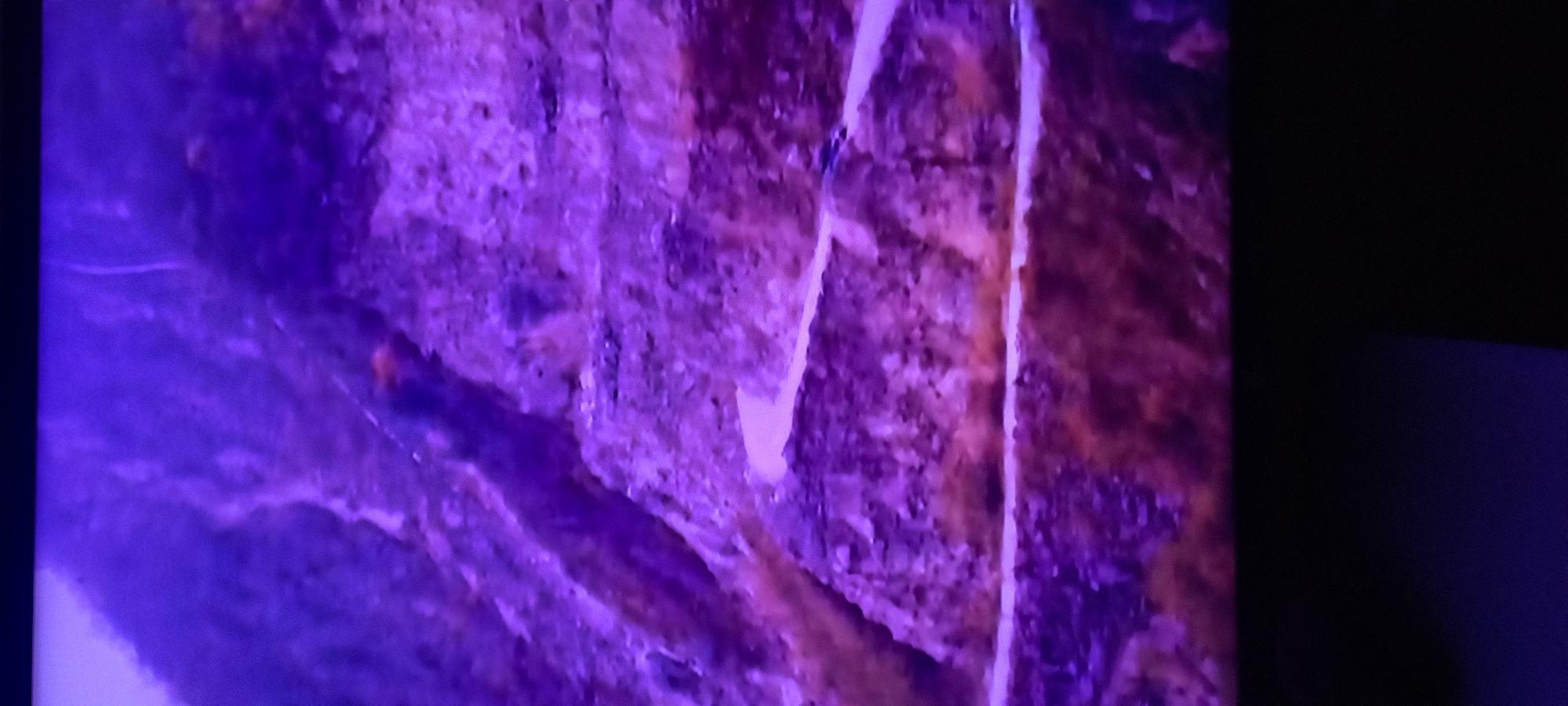 An aerial perspective captures a steep, mountainous landscape bathed in a vibrant purple hue. A winding road carves its way through the terrain, appearing as a pale ribbon against the rugged, textured mountainside. Small, dark shapes, likely vehicles, can be seen traversing the road. The overall impression is one of a remote and dramatic natural setting, possibly a mountain pass or a remote road in a challenging environment.