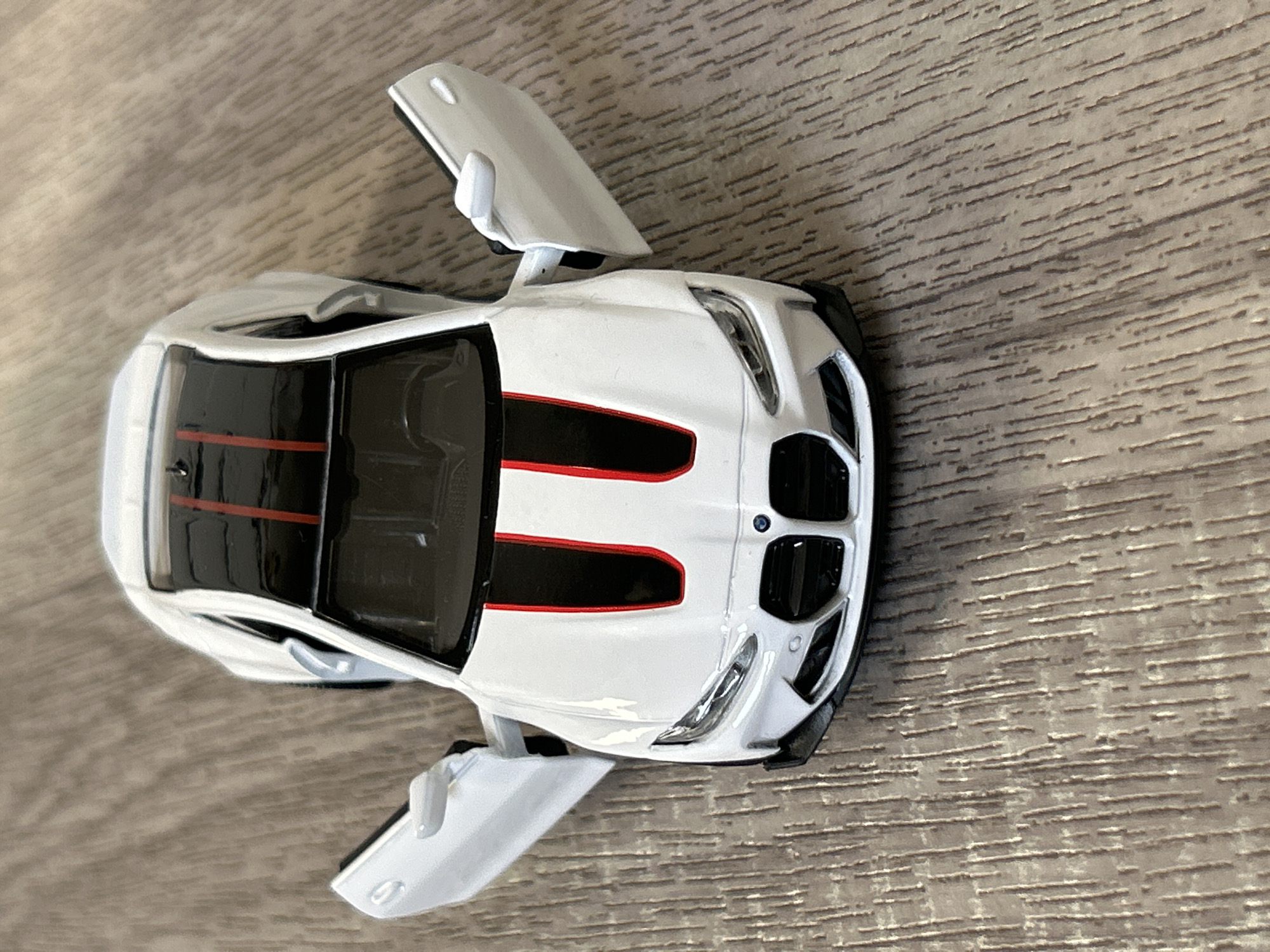 A white toy BMW with red racing stripes on the hood and roof is centered on a horizontally oriented faux wood surface. The doors are open upward, and the interior of the car is black.