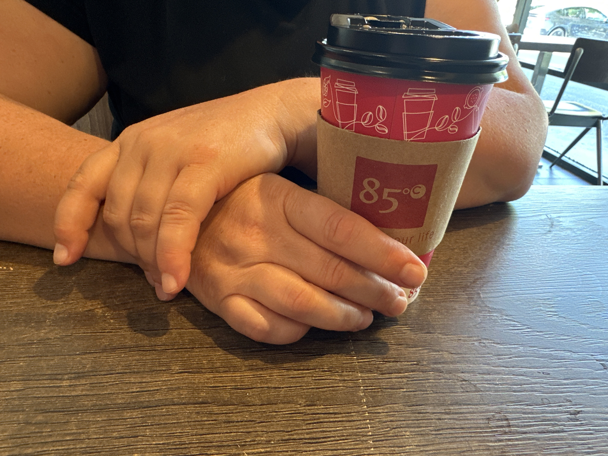 A close-up shot shows a person's hands resting on a wooden table in what appears to be a coffee shop in Lynnwood, United States of America. In the center of the frame is a disposable coffee cup with a red sleeve adorned with white line drawings of coffee cups and beans, and the number "85°" in white font. The cup has a black lid. The person's arms and hands are positioned around the cup, with one hand resting on top of the other, fingers gently interlocked. The background is blurred, but hints of a window, chairs, and a vehicle can be seen, suggesting an indoor setting with a view of the outside. The lighting is natural and casts soft shadows on the table.
