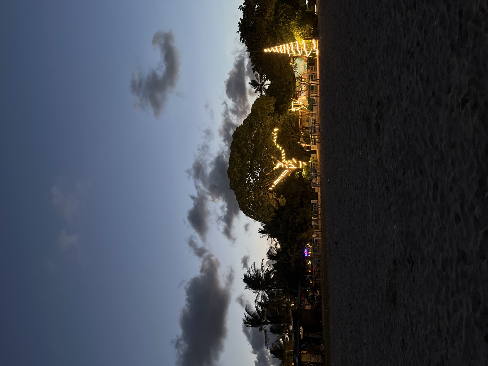 The image captures a scene at sunset on a beach in Kuta Selatan, Indonesia. The sky is a gradient of blue and grey, with scattered, dark clouds. Along the beach, there are palm trees and larger trees adorned with decorative string lights. Some of these trees are arranged with seating underneath, suggesting a beachside dining or recreational area. The ground is covered with dark sand. The scene appears to be devoid of people.