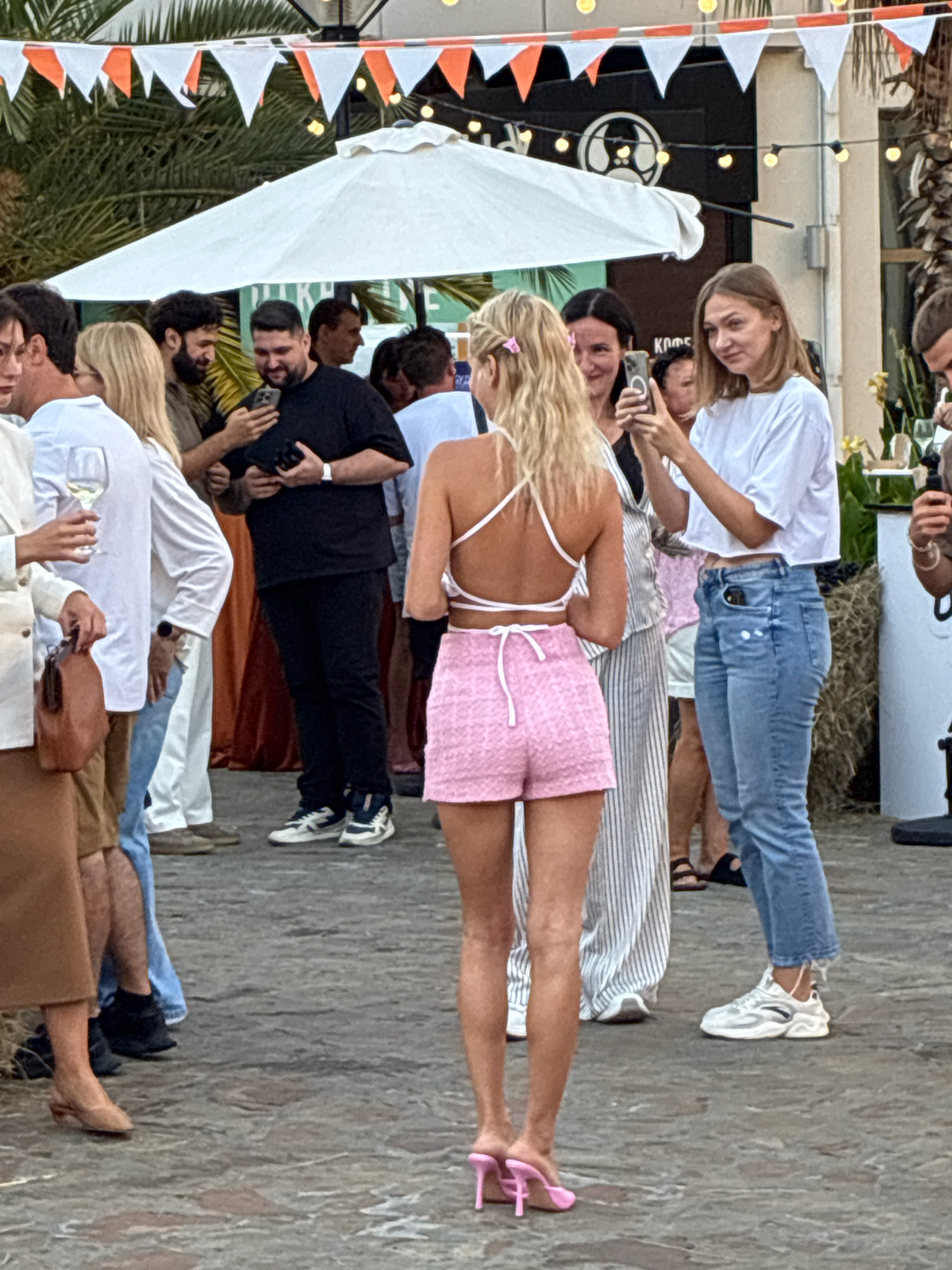This is a medium shot, taken from a slightly low angle, of a group of people outdoors in what appears to be a public gathering area, possibly a resort or festival, in Sochi, Russia. The background features palm trees and a building with decorative orange and white triangular flags strung across the top, typical of a festive atmosphere.
In the foreground, the back of a woman with blonde hair is visible. She is wearing a white halter-neck crop top and pink textured shorts, paired with pink heeled mules. She is looking to her right, away from the camera.
To her left, a woman with dark hair is smiling and holding a smartphone, seemingly taking a picture or recording. She is wearing a striped dress. Next to her, a man in a black t-shirt and black pants is looking down at his phone, holding what appears to be a wallet in his other hand. He is wearing white sneakers with black accents.
Further left, a woman in a white blazer and brown skirt is holding a wine glass. Behind her, a man with dark, curly hair is looking at his phone. Another man in a white shirt and jeans is visible behind them.
The ground is paved with cobblestones. The overall scene suggests a lively outdoor event.