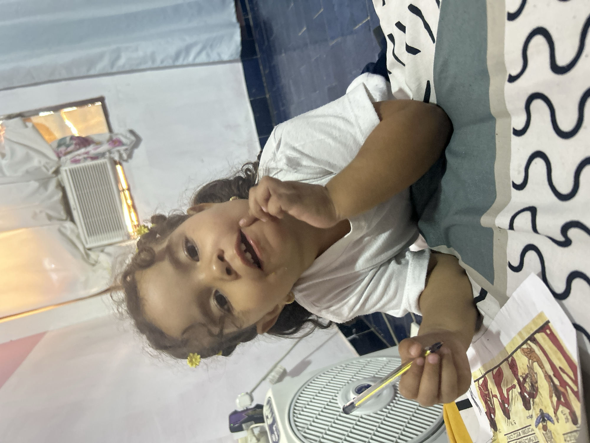 A young child with curly hair and yellow flower decorations in their hair is lying on a bed with a patterned comforter. The child is holding a pen in their right hand and has their left hand near their mouth. In the foreground, there is a white fan and a piece of paper with an Egyptian-themed drawing on it. In the background, a window with closed blinds and a small air conditioning unit can be seen. The location is a room in a house, and given the prompt, it is presumably located in San Juan de Los Morros, Venezuela.