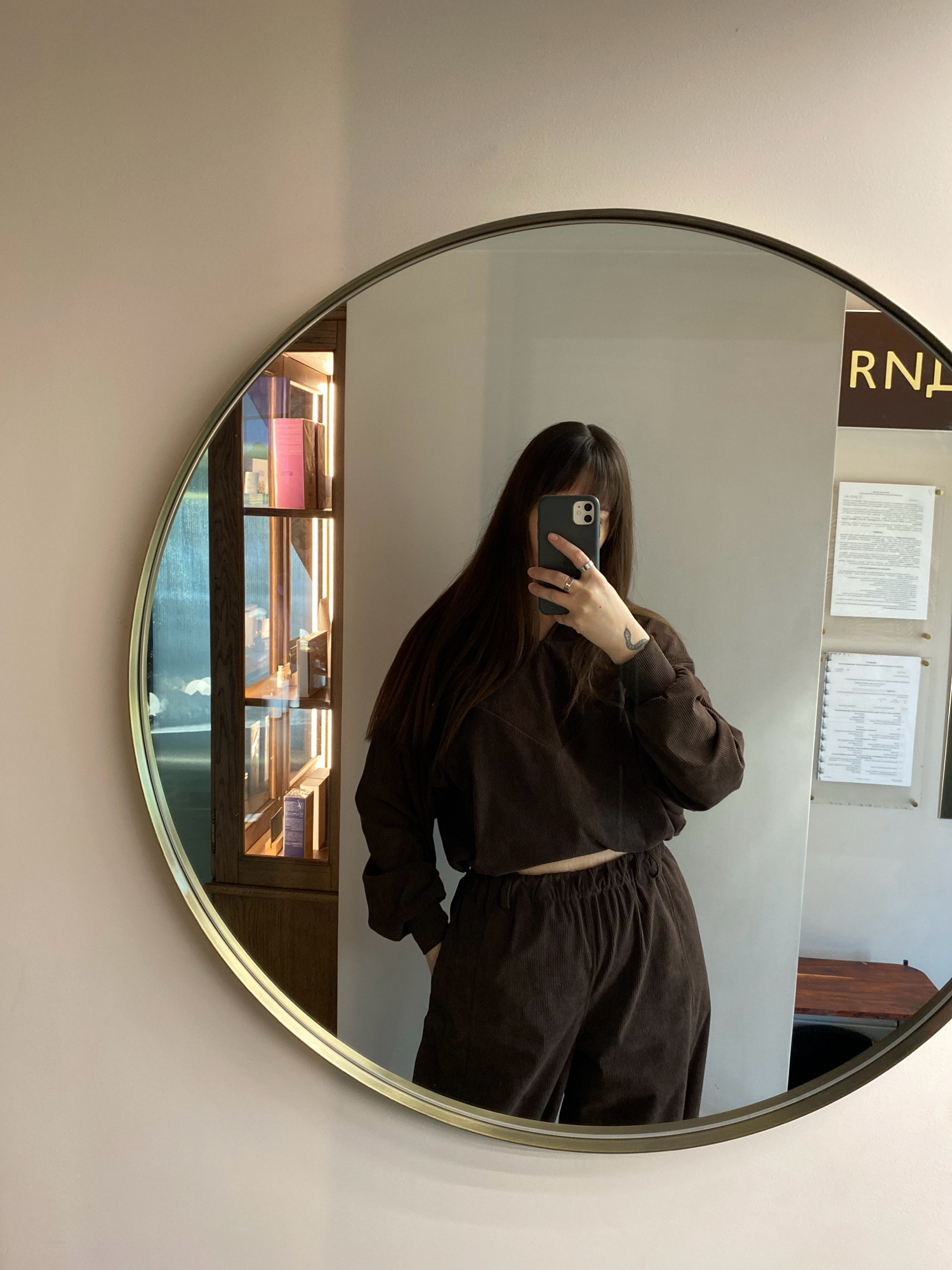 The image shows a woman taking a mirror selfie in a room with a light-colored wall. She is wearing a brown corduroy two-piece set, consisting of a cropped long-sleeved top and pants. Her dark hair falls over her shoulders, and she is holding a smartphone in her right hand. A circular mirror with a thin gold frame reflects her image and part of the room. To the left of the woman, visible in the mirror, is a tall, narrow wooden cabinet with glass doors, illuminated from within, containing several products on shelves. To the right of the woman, also visible in the mirror, are two documents affixed to a light-colored wall, and a portion of a dark wooden surface. The overall lighting suggests it may be daytime. The location is not discernible from the image.