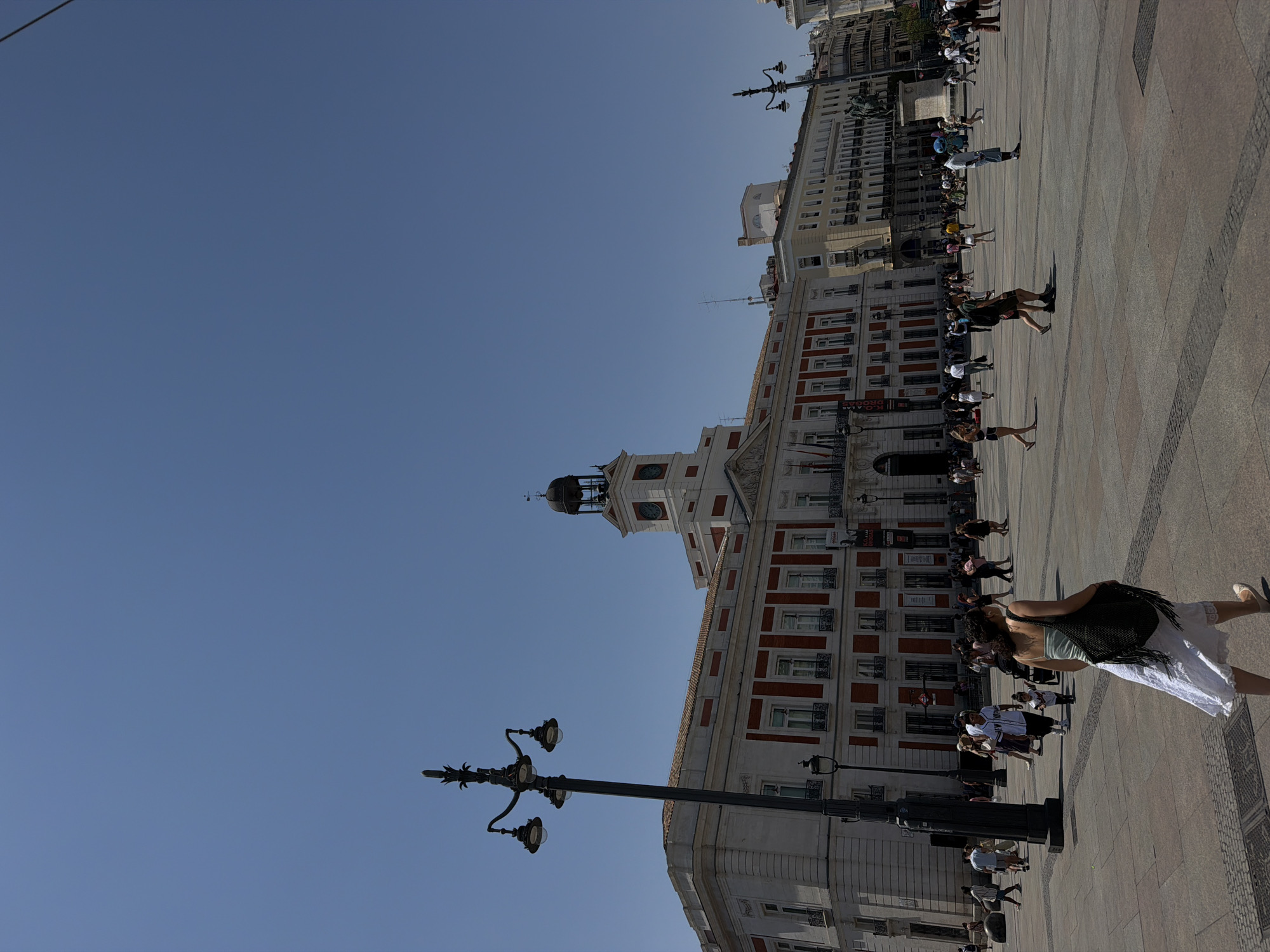The image shows a wide-angle view of the Puerta del Sol in Madrid, Spain, on a clear, sunny day. The iconic Real Casa de Correos building with its clock tower dominates the background, featuring a white facade with red accents and numerous windows. A tall, ornate black lamppost with three lamps stands in the left foreground. The plaza is paved with large stone tiles and cobblestones. Many people are walking across the plaza, some individually, some in small groups. Some are dressed in casual summer clothing. In the foreground, a woman wearing a white skirt and a light-colored top with a dark, tasseled bag slung over her shoulder walks away from the camera. The sky is a clear, bright blue with no clouds. The overall impression is a bustling public square under a bright summer sun.