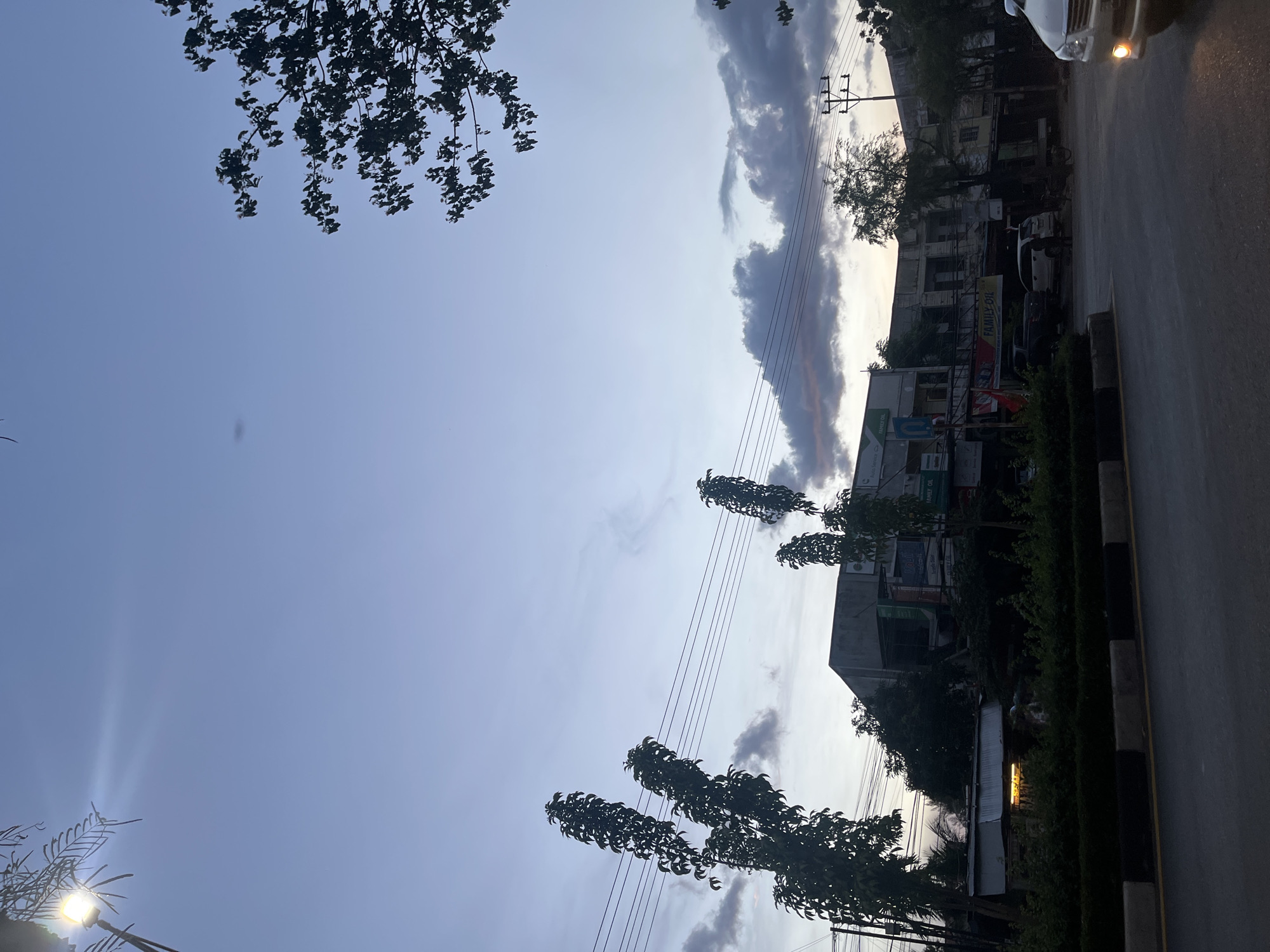 The image shows a street scene in Banda Aceh, Indonesia, during twilight. A street lamp in the upper left corner emits a bright beam of light. In the foreground, a road is visible with a median strip. Beyond the road, there are several buildings that appear to be shops or businesses, with some trees and power lines in front of them. A few vehicles are parked or driving on the street. The sky is a pale blue with some scattered clouds, suggesting either dawn or dusk. The overall impression is of a quiet, urban environment at the end of the day.