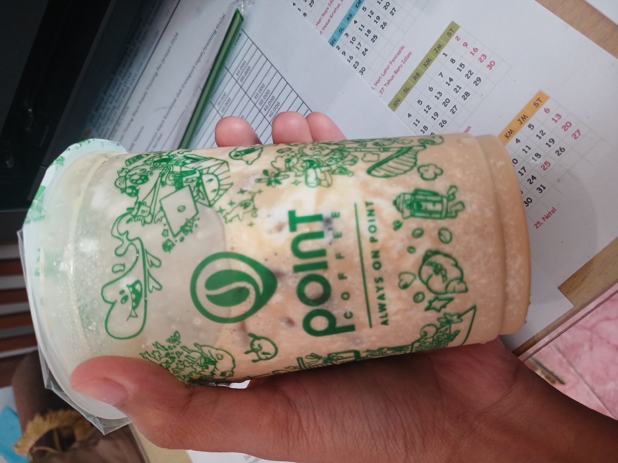 A hand holds a plastic cup of iced coffee from "Point Coffee" with green cartoon illustrations and branding. The cup is filled with a beige, frothy liquid. In the background, a calendar page shows dates in March. The scene appears to be in an indoor setting, possibly an office, with papers and a printer visible. There is no discernible information about the city of Watugede, Indonesia, within the image.