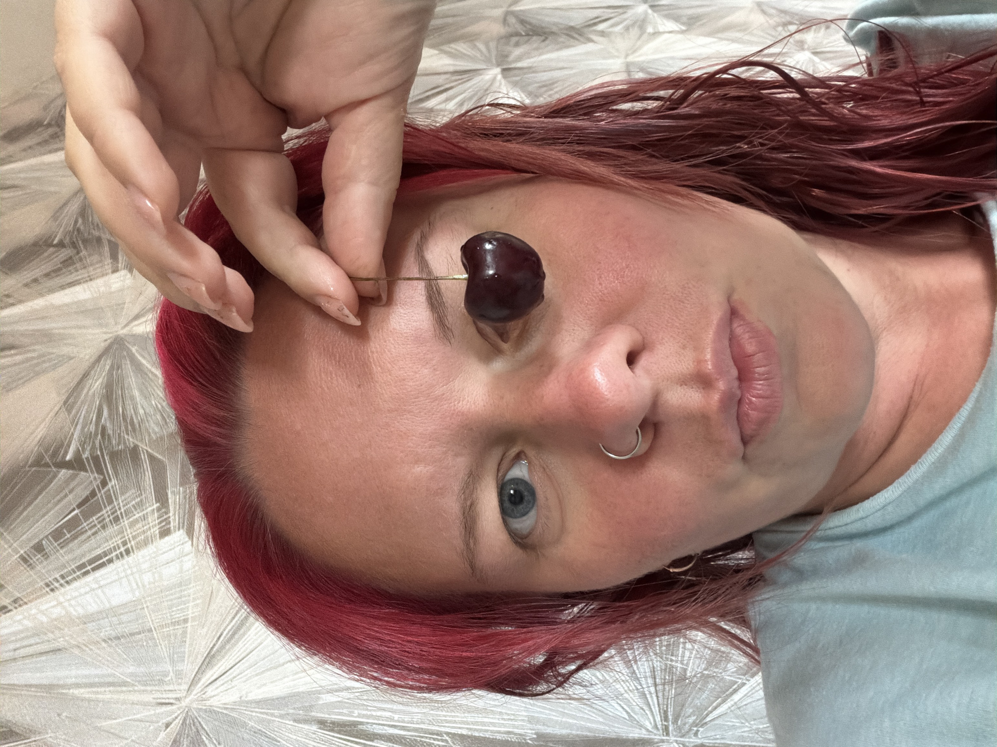 The image shows a close-up of a person's face with bright red hair. The person is holding a cherry with their right hand, placing it over their right eye. They are wearing a light blue t-shirt and have a nose ring. The background appears to be a textured wallpaper with a geometric pattern. The lighting suggests an indoor setting. There is no discernible information within the image that would allow for the identification of a specific location in the city of Chaikovsky, Russia.