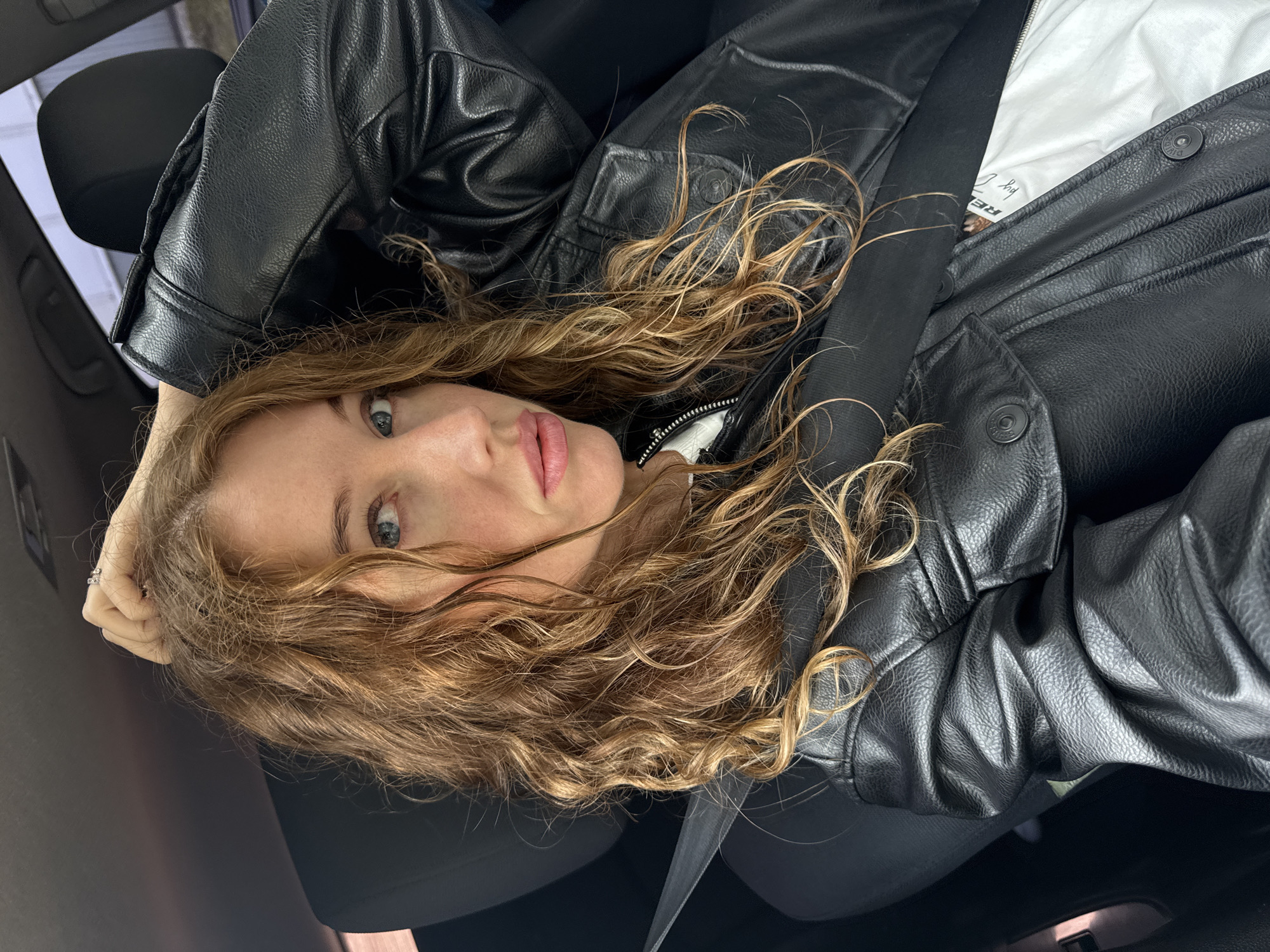 The image depicts a woman seated in the passenger side of a vehicle in Chaikovsky, Russia. The woman has wavy, reddish-brown hair with lighter highlights, blue eyes, and light skin with visible freckles or moles. She is wearing a black leather jacket over a white t-shirt, a portion of which reveals a white label with "REPLAY" text. A dark seatbelt crosses her torso and lap. Her right hand, adorned with two rings on the ring finger, is positioned near the top of her head, resting in her hair. Her left hand is not fully visible. The interior of the car is dark, featuring black seats and dark upholstery. Through the car window in the upper right, a muted view of an exterior structure or building is visible under an overcast sky. The woman is looking directly at the camera with a neutral expression and slightly parted lips.