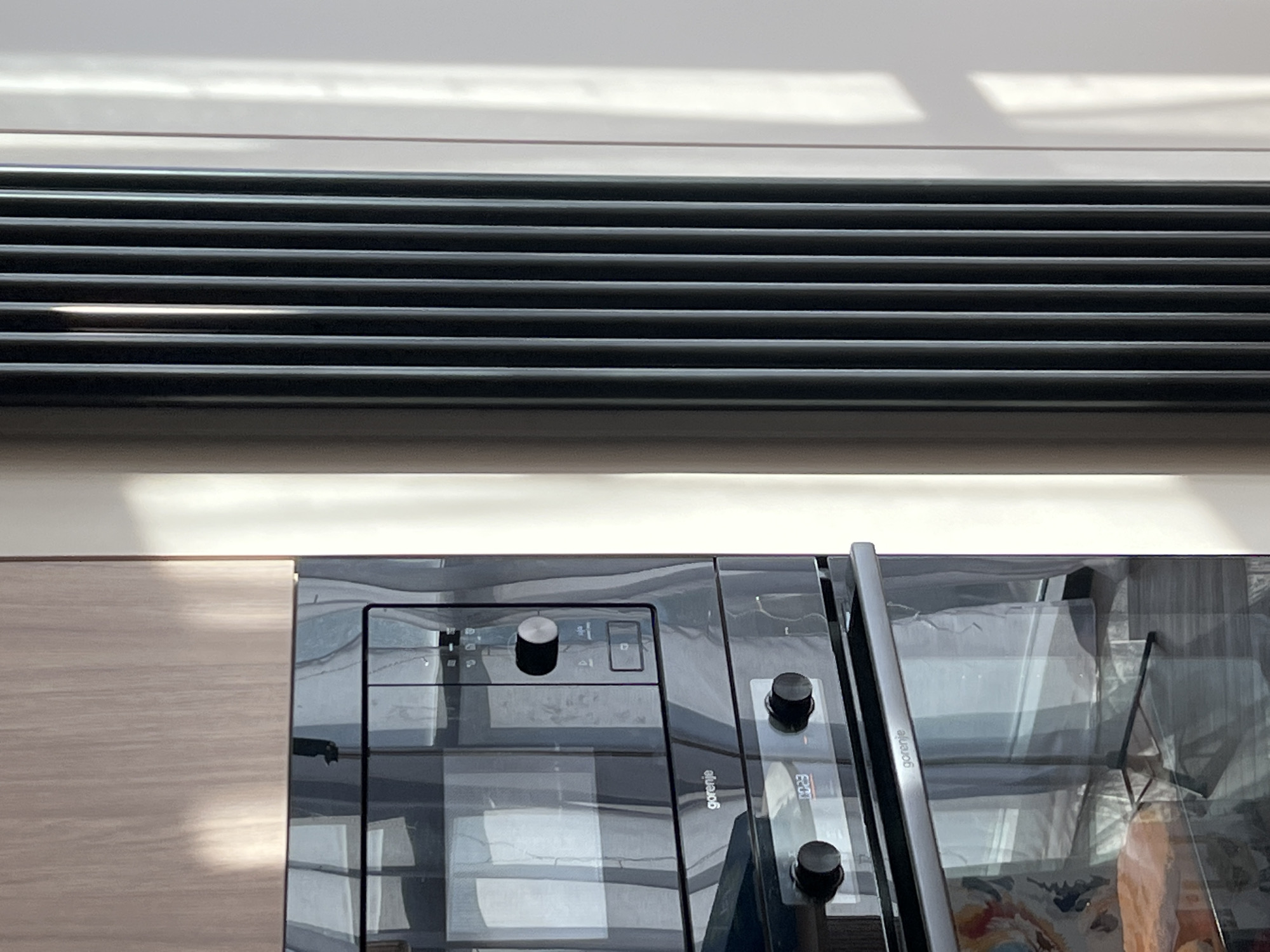 The image shows a section of a kitchen interior. A horizontal black air vent is located near the top of the frame. Below the vent is a beige cabinet and a black oven/microwave appliance with the brand name "Gorenje" visible on its surface. The environment appears to be indoors, and the light source is coming from above. The appliances and cabinets suggest a residential kitchen setting. There is no information available about the location in the city of Новое, Russia.