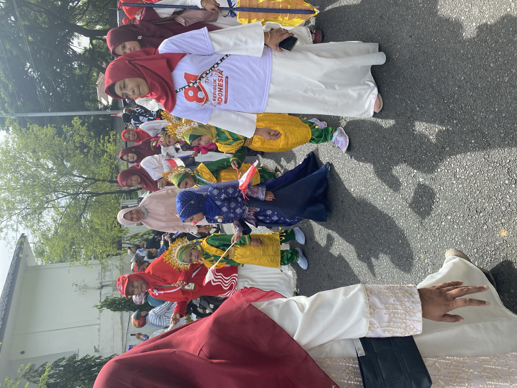 A street parade is taking place in Banda Aceh, Indonesia, as indicated by the Indonesian flag and festive attire. A group of people, predominantly women wearing hijabs, are walking in a procession on an asphalt road. Several children are also present, dressed in traditional and colorful outfits. Some of the children are holding small Indonesian flags, symbolizing national pride. In the background, there are buildings and trees, suggesting an urban setting. The overall atmosphere appears to be celebratory and communal, likely for a national holiday or event.