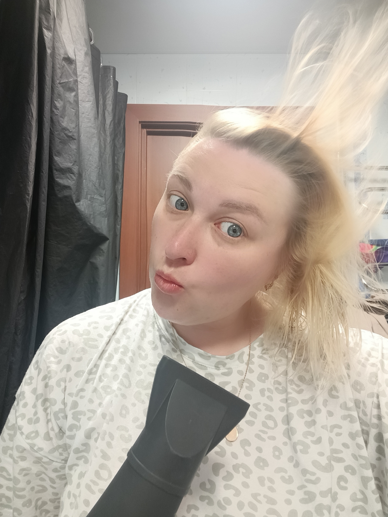 A woman is holding a black hair dryer and drying her blond hair in what appears to be a bathroom. She is wearing a white, animal-print t-shirt and is looking at the camera with her lips pursed. A black shower curtain is visible on the left, and a brown door is in the background. The location is not evident from the image.