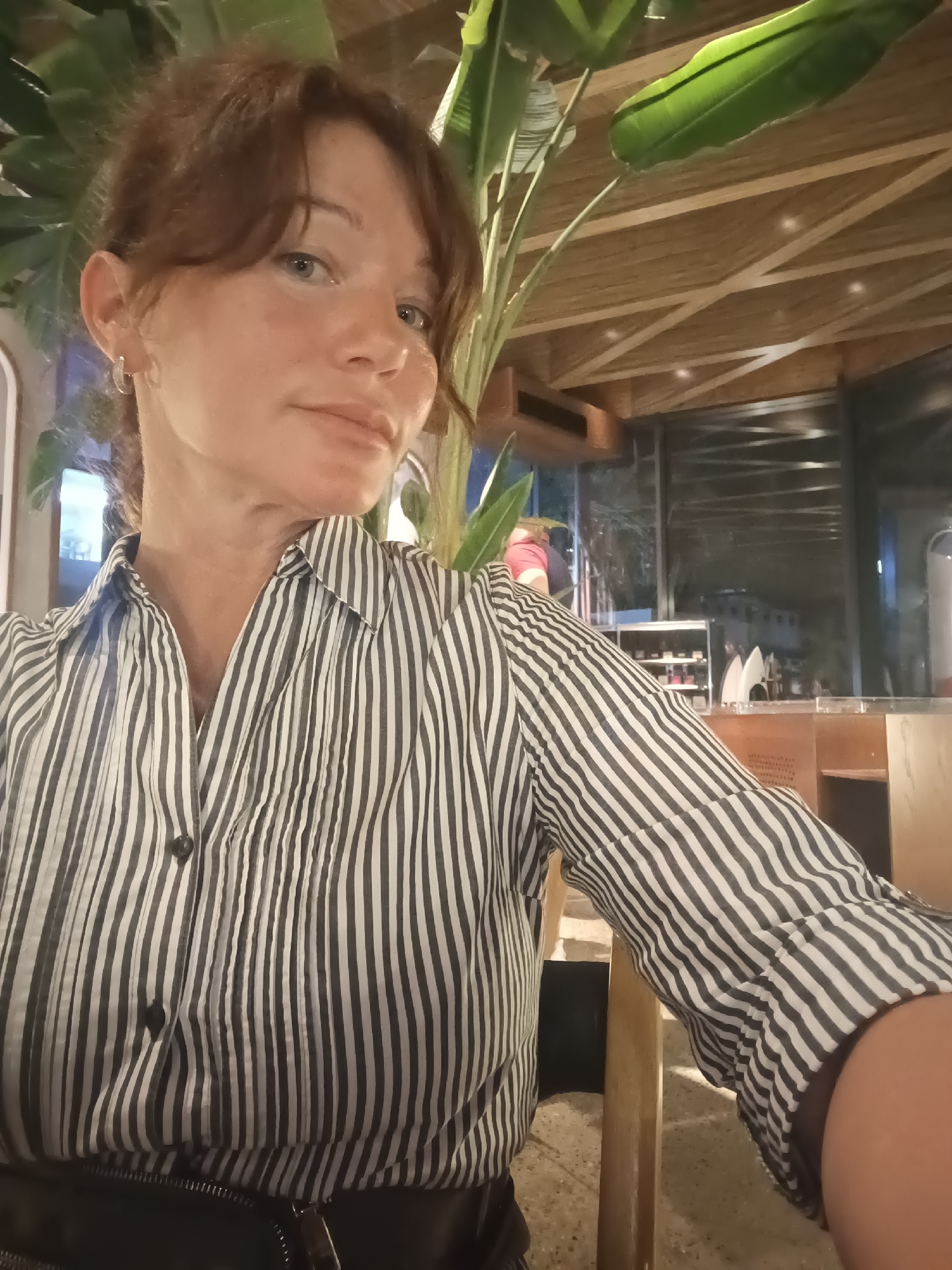 A woman with auburn hair tied back is seated at a table in an indoor setting with a thatched ceiling and bamboo accents. She is wearing a black and white striped button-up shirt and a black belt or waistband is visible at the bottom of the frame. Large green leaves from a plant are in the background. The overall ambiance suggests a restaurant or bar. The lighting is dim, with some spotlights on the ceiling and ambient light from the exterior visible through a large window or glass partition in the background. The woman is looking towards the camera and her expression is neutral.