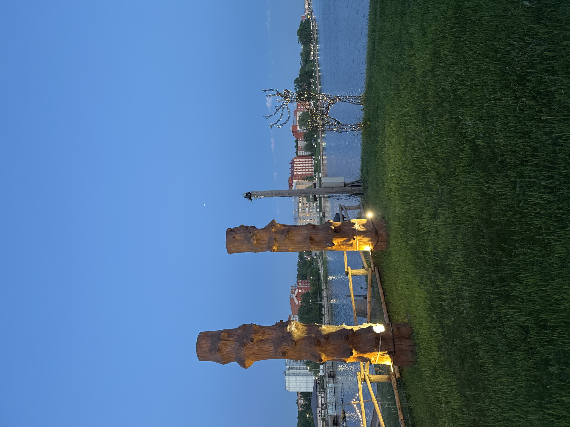 The image shows an evening scene in Votkinsk, Russia. In the foreground are two upright log structures, illuminated by warm lights at their bases. They are connected by a simple wooden frame. Behind them is a view of the city's waterfront along the Votkinsk Reservoir. A light-up deer figure stands on the green lawn, further back towards the water. Buildings are visible across the reservoir. The sky is a clear, deep blue.