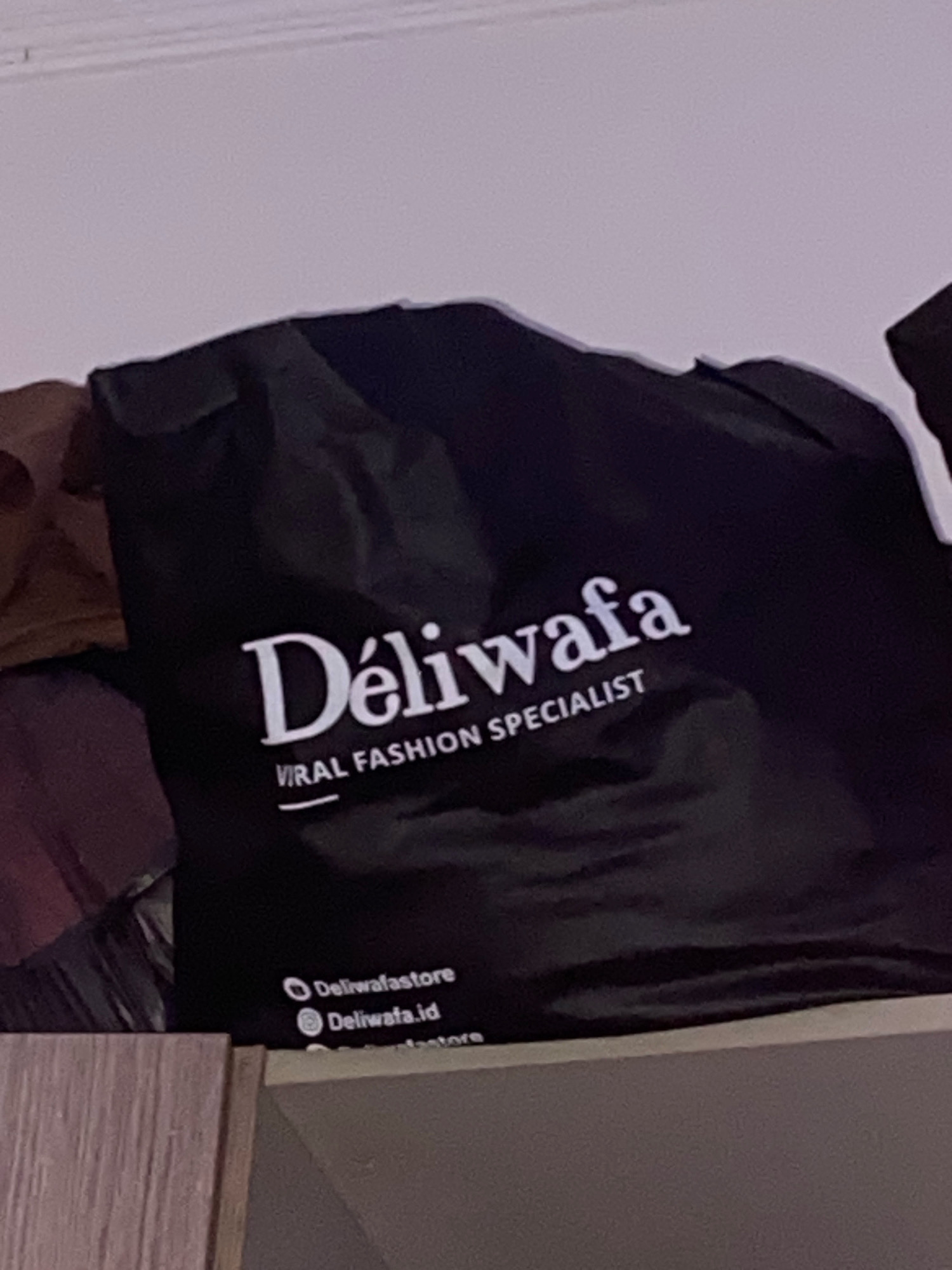 In the city of Klurak, Indonesia, a black fabric bag is prominently displayed, possibly on a shelf or within a wardrobe, as indicated by the wooden surface below. The bag is adorned with white text, featuring the brand name "Déliwafa" in a stylized font, followed by the tagline "VIRAL FASHION SPECIALIST." Below this, two social media handles are visible: "Deliwastore" and "Deliwafa.id," each preceded by a small circular icon. The background is a plain, light-colored surface, likely a wall or ceiling. The lighting appears somewhat dim, casting subtle shadows and highlighting the textures of the fabric.