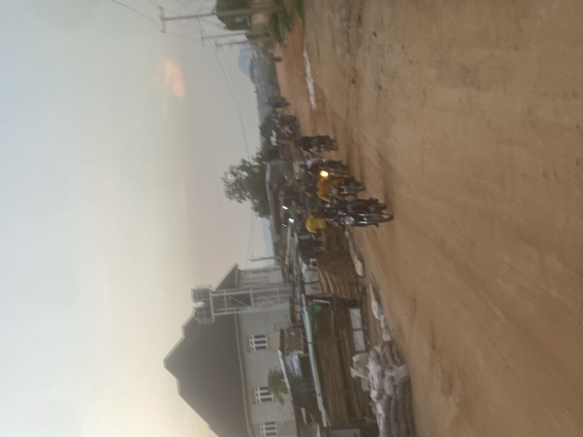 The image depicts a semi-urban scene in Lugbe, Nigeria, under what appears to be late afternoon or early evening lighting conditions. The foreground and mid-ground are dominated by an unpaved, reddish-brown dirt road that shows evidence of vehicle tracks.
On the left side of the road, a large, two-story building with light-colored walls and a dark, multi-gabled roof is prominent. Adjacent to this building is a tall, metallic structural tower, supporting two water tanks at its apex. In front of the building and along the roadside, there are stacks of raw lumber or planks of wood and several white sacks, likely containing cement or other construction materials. Other smaller, less defined structures are visible in the vicinity of the main building.
The dirt road is occupied by a procession of at least seven motorcycles, commonly known as okadas, moving generally from the background towards the viewer's left, but angled to move through the scene. Each motorcycle has at least one rider; some appear to carry passengers. One rider in the mid-ground wears a distinct yellow shirt, while another behind him is dressed in a yellow-brown garment. The headlights of some motorcycles are illuminated.
On the right side of the image, beyond the dirt road, utility poles with power lines are visible, extending into the distance. The background features hazy distant hills or mountains under a pale, diffused sky that exhibits a faint orange hue in one section, consistent with either dawn or dusk. The overall impression is one of ongoing development or a busy local thoroughfare within a developing community.