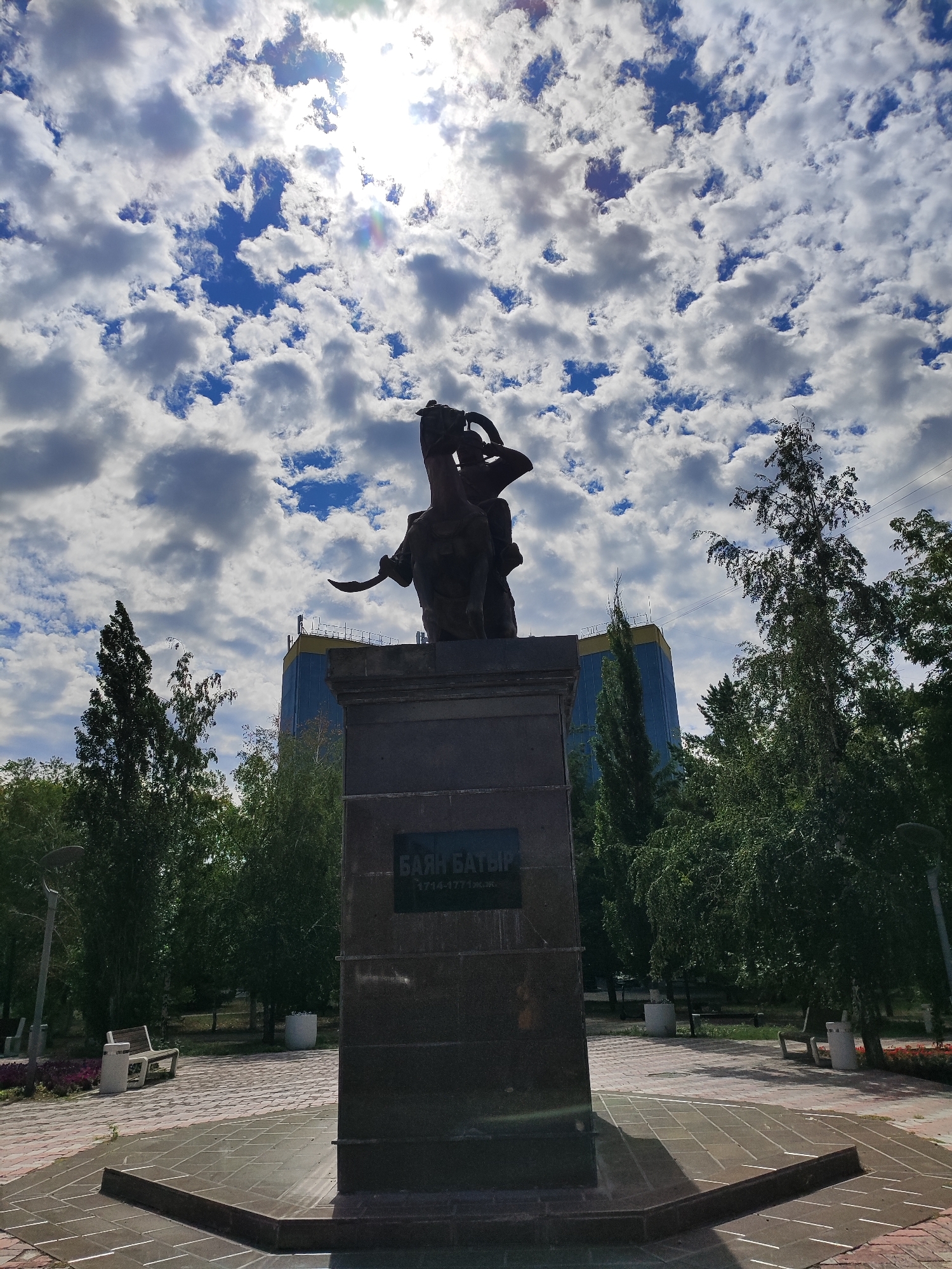 The image shows a monument to Baian Batyr in Pavlodar, Kazakhstan, under a partly cloudy sky. The monument, made of dark stone, features a statue of a warrior on horseback. The base of the monument has an inscription that reads "БАЯН БАТЫР 1714-1771 гг." The monument is located in a park area with trees, benches, and a paved walkway. In the background, a modern building with blue and yellow facades is visible. The sun is shining brightly through the clouds, creating lens flare.