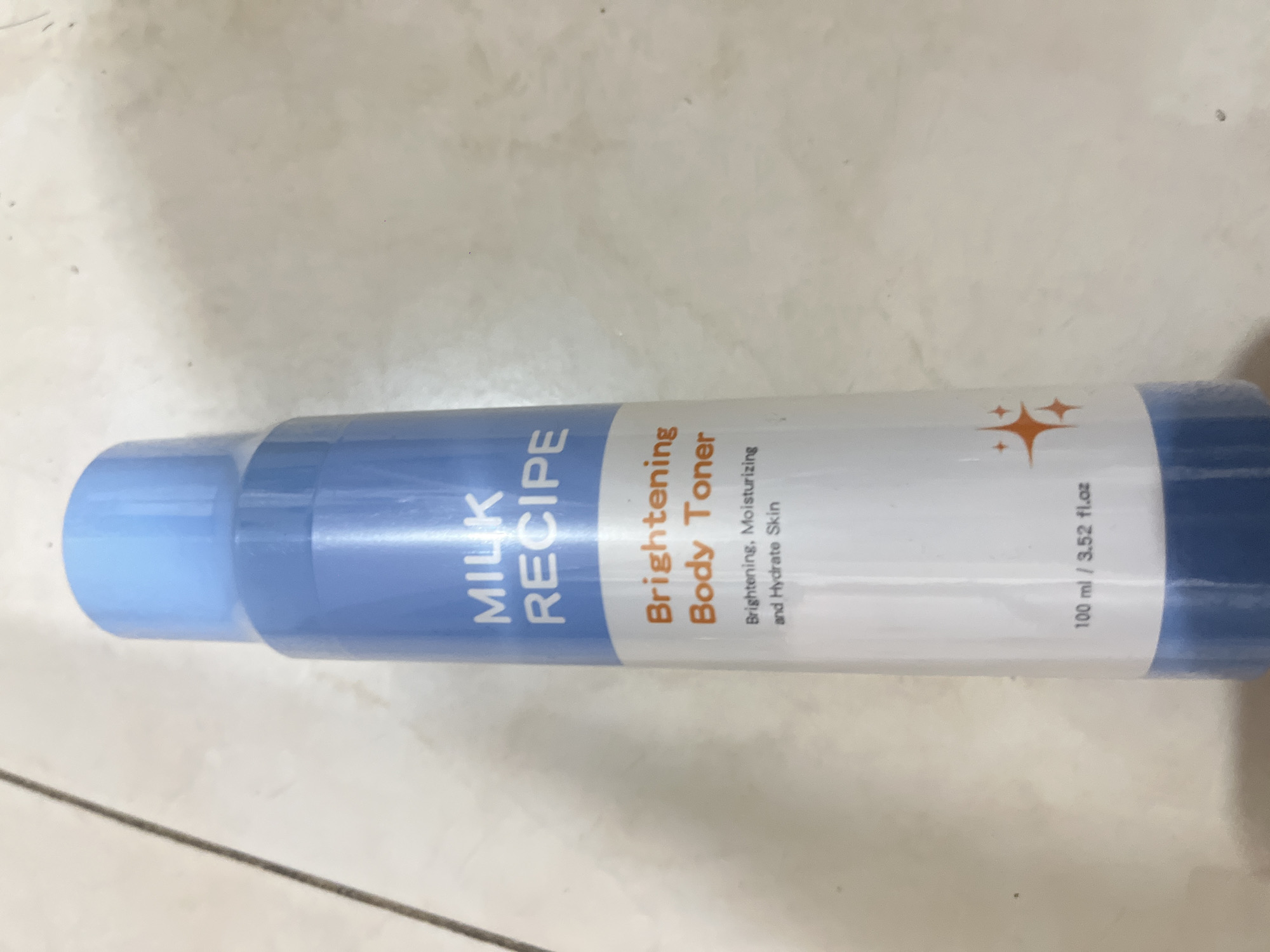 A tall, slender, light blue plastic bottle stands upright on a light-colored, tiled surface. The bottle has a white label with blue and orange text. The text on the label reads "MILK RECIPE," "Brightening Body Toner," "Brightening, Moisturizing and Hydrate Skin," and "100 ml / 3.52 fl.oz." Two small, orange starburst graphics are printed on the lower part of the label. The background is a textured, light-colored surface, possibly a wall or floor, with faint shadows. The scene is indoors, with no indication of its specific location within Denpasar, Indonesia.
