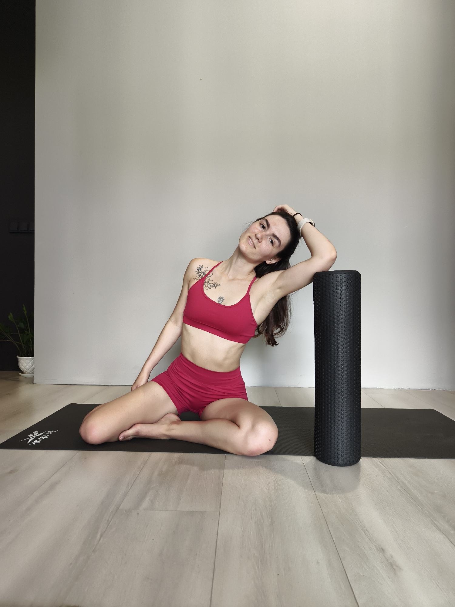 An indoor, eye-level shot from Tyumen, Russia, captures a woman engaged in a stretching exercise. She is seated cross-legged on a black yoga mat, facing the viewer with her head tilted to her left. Her right arm is bent with her hand behind her head, and her right elbow rests on a tall, black, textured foam roller that stands upright beside her.
The woman has dark hair and wears a red sports bra and matching red athletic shorts. Floral tattoos are visible on her left shoulder and chest. She has a subtle, calm expression on her face as she looks towards the camera.
The background consists of a plain light grey wall, with a darker grey or black section and a small potted plant visible on the far left. The floor is light-colored wooden planks. The lighting is bright and even, suggesting a daytime scene. A white "Reebok" logo is clearly visible on the black yoga mat. The overall scene conveys a sense of peaceful wellness and focused activity.