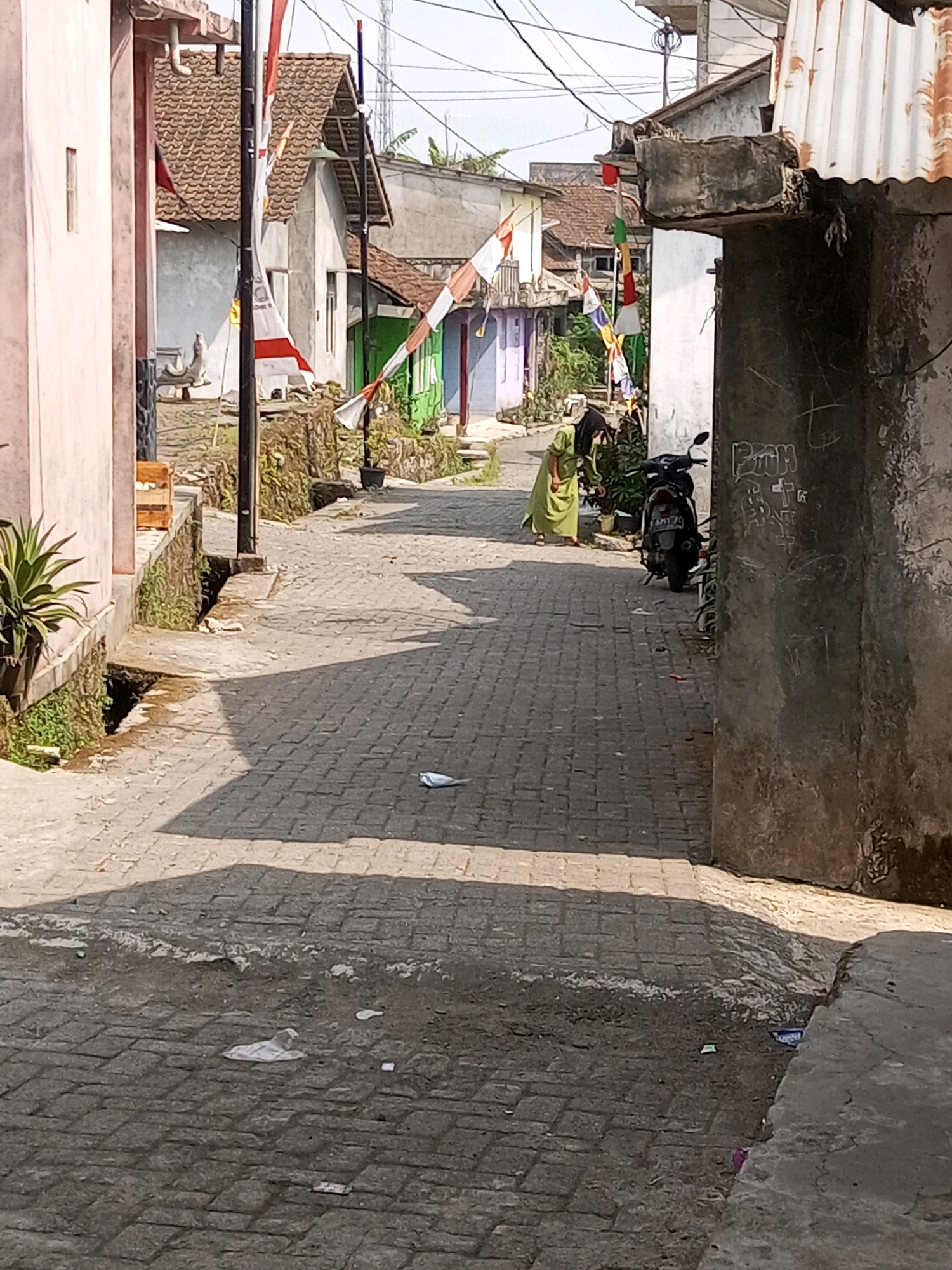 The image depicts a narrow, cobblestone street in Andongsili, Indonesia, flanked by modest residential buildings. The houses feature tiled roofs, some with a rustic, weathered appearance. Colorful flags, likely for a celebration, are strung across the street, adding a festive touch to the scene.
In the middle ground, a person wearing a green hijab and traditional clothing walks away from the camera, towards the right side of the street. A dark-colored motorcycle is parked on the right side of the road, near the person. The street surface is paved with interlocking cobblestones and shows signs of wear and tear. Shadows cast by the buildings and the flags create a dappled pattern on the ground.
Various items of litter are scattered on the street, including a crumpled white object that appears to be a face mask in the foreground, and other small pieces of debris further down the road. The environment appears to be an ordinary residential area, possibly a village or a less developed part of a town. The overall impression is one of a warm, sunny day in a community setting.