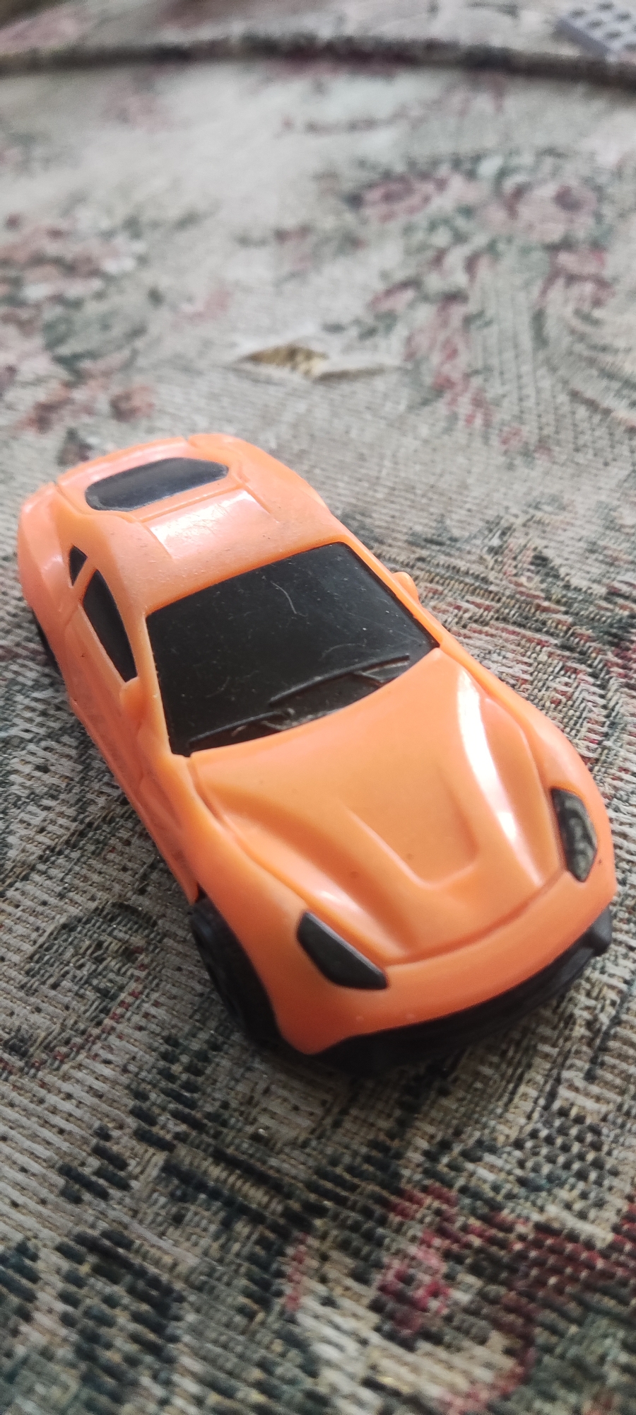 An orange toy sports car sits on a patterned fabric surface. The car is a miniature replica of a sleek, modern vehicle, with black tinted windows and black wheels. The fabric beneath it has a floral design in shades of pink, green, and grey. The image appears to be a close-up shot, with the car occupying the majority of the frame. The lighting suggests an indoor environment, possibly a home. There are no people or other objects clearly visible in the scene. The location is identified as Gebang, Indonesia, though there are no visual cues in the image to corroborate this.