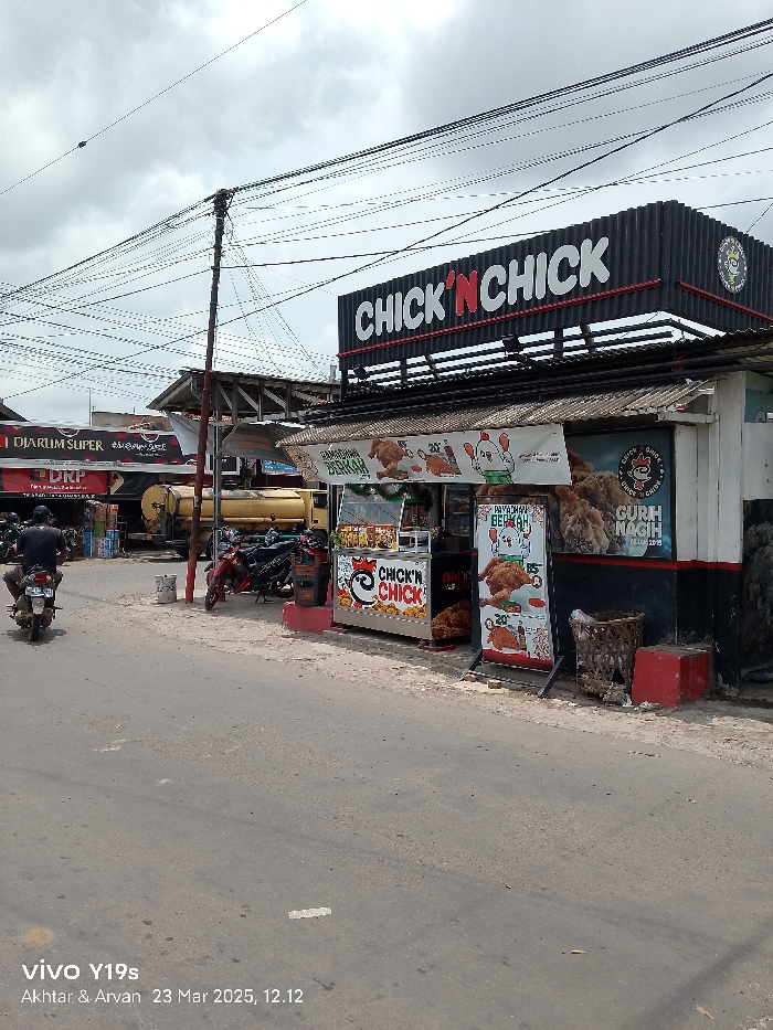 The image shows a street scene in Kecamatan Haurgeulis, Indonesia. A small food stall with a sign that reads "Chick 'n Chick" in red and white letters is prominently featured. A person riding a motorcycle is driving down the street. The motorcycle is parked near a truck and a stand with several motorcycles parked. There are power lines overhead, and the sky is partially clouded. There are several colorful signs depicting fried chicken and other food items. The stall's walls are black and adorned with numerous additional signs. The stall's roof is made of corrugated metal. The pavement is gray and weathered. There are some plants and a trash bin outside the stall.