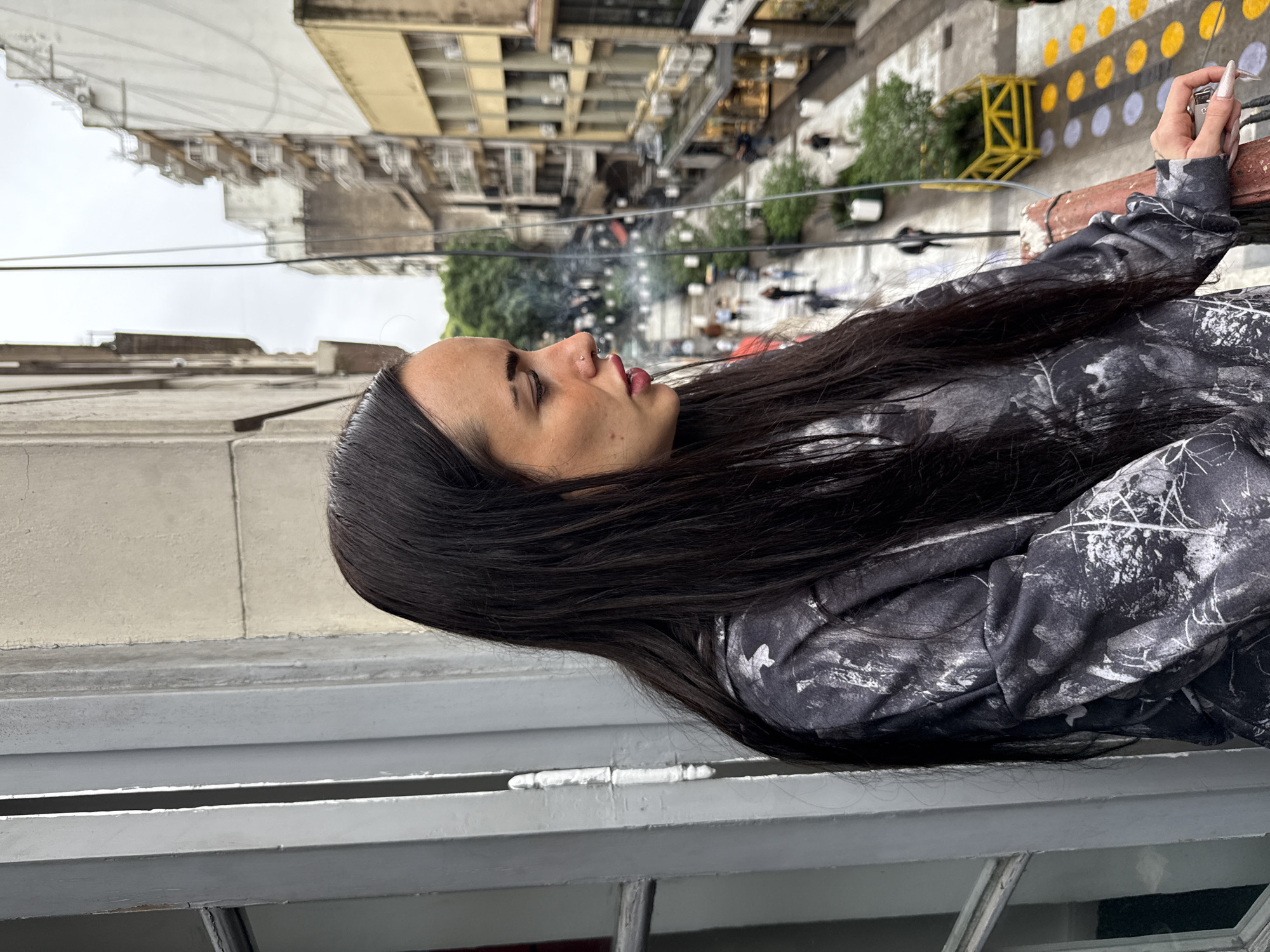A side-profile shot captures a young woman with long, dark hair, looking out from a window or balcony over a city street. She is positioned on the right, facing left and slightly upwards. Her lips are slightly parted, and she has a small piercing on her nose. She wears a dark long-sleeved top or jacket featuring a white and grey abstract or camouflage-like pattern. Her right hand, adorned with long, light-colored fingernails, holds onto a dark railing and clutches a small, metallic silver-colored object, possibly a lighter or vape device.
In the foreground, a light grey painted metal window frame or balcony railing is visible. The background reveals an urban cityscape under an overcast, light grey sky. Numerous buildings of varying heights and architectural styles, predominantly in light beige and grey tones, line the street. Below, the street level shows pedestrians, green trees, and street furniture. Notable details in the middle distance include a wall or ground structure decorated with distinctive yellow and white circular patterns, alongside some yellow metal structures. The scene suggests a contemplative moment on a cloudy day in Buenos Aires, Argentina. No discernible text is visible in the image.