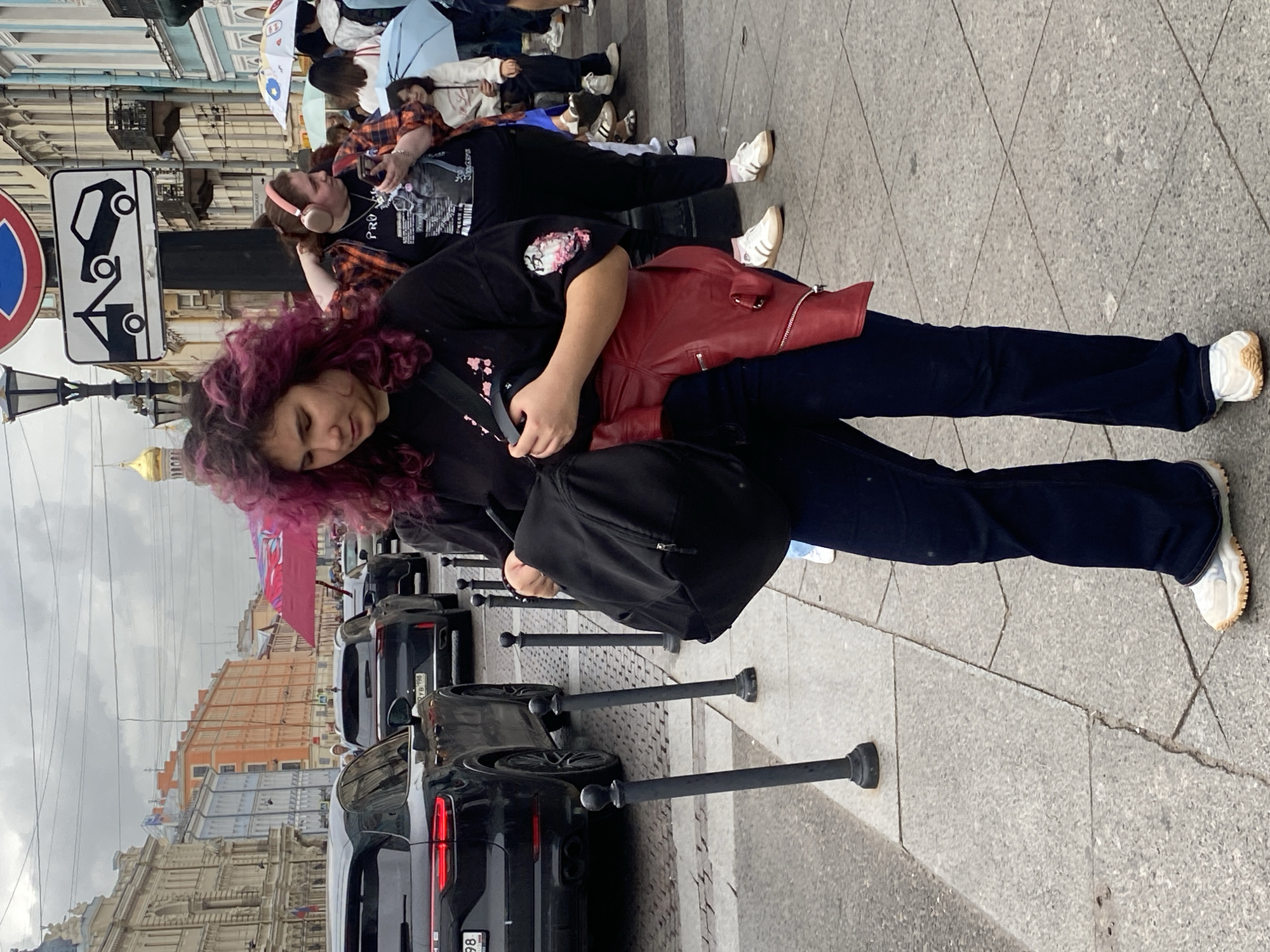 The image captures a street scene in Saint Petersburg, Russia, under an overcast sky. In the foreground, a young woman with distinctive purple-dyed hair is holding a black backpack and appears to be adjusting something within it. She is wearing a black t-shirt, dark flared jeans, and white sneakers with tan soles. A red leather jacket is draped over her waist. Behind her, another woman with pink headphones is looking at her phone. Several other people, some holding umbrellas, are visible in the background, along with a line of cars, including a black Porsche SUV. The street is paved with cobblestones, and bollards line the sidewalk. Buildings with varied architectural styles, typical of Saint Petersburg, are seen along the street. A tow-away zone sign is also visible.