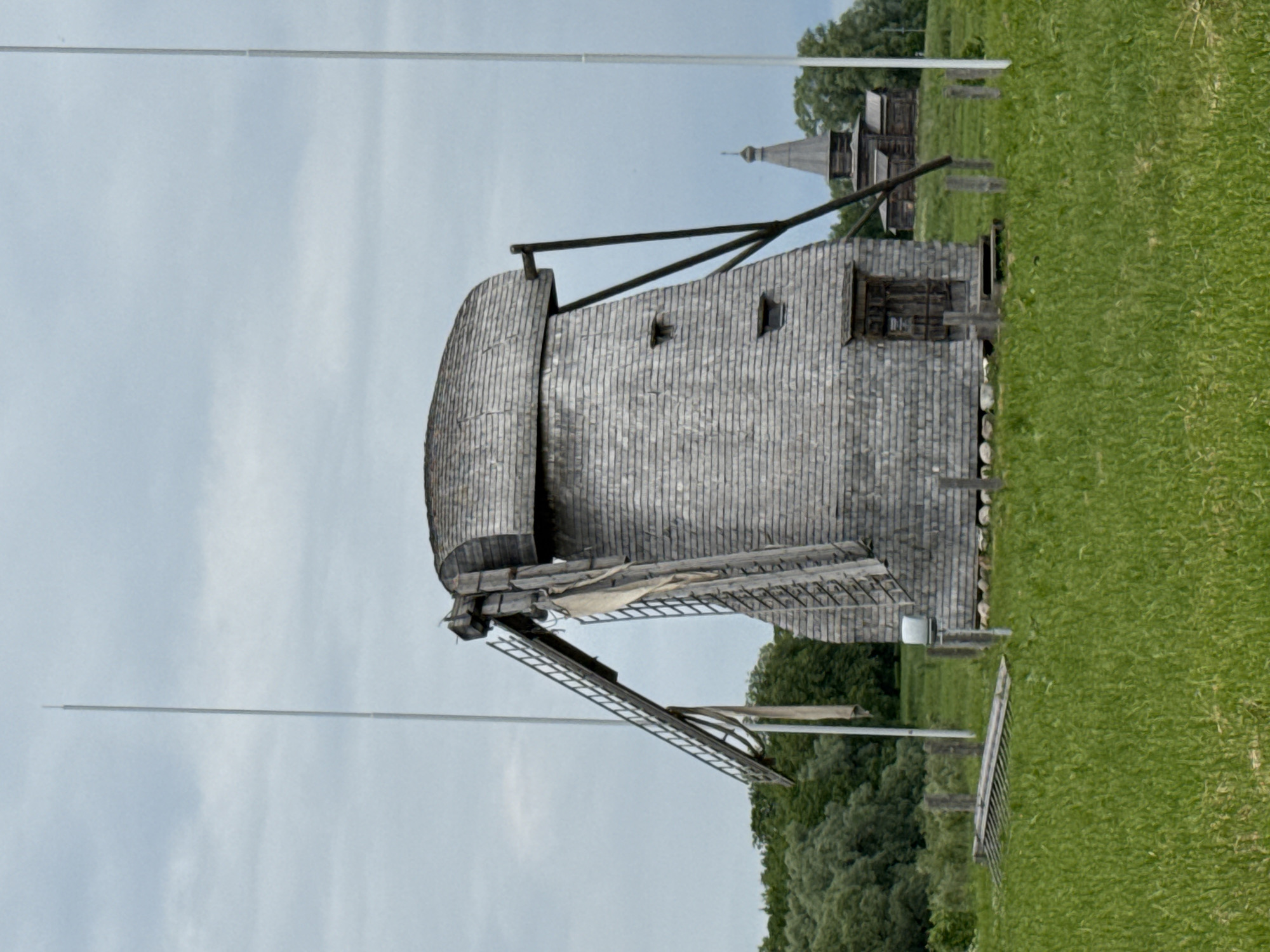 A traditional wooden windmill stands in a grassy field under a cloudy sky. The windmill, covered in weathered grey wooden shingles, has a distinctive curved roof and a ladder leading up to its side. Two tall white poles are visible on either side of the windmill, with a portion of a wooden structure resembling a church in the background, behind a line of trees. The grass in the foreground is lush and green, with a few wooden posts and a section of a wooden structure lying on its side. The image appears to be taken at the Vitoslavlitsy Museum of Folk Wooden Architecture located on the shores of Lake Myachino, near Veliky Novgorod, Russia.