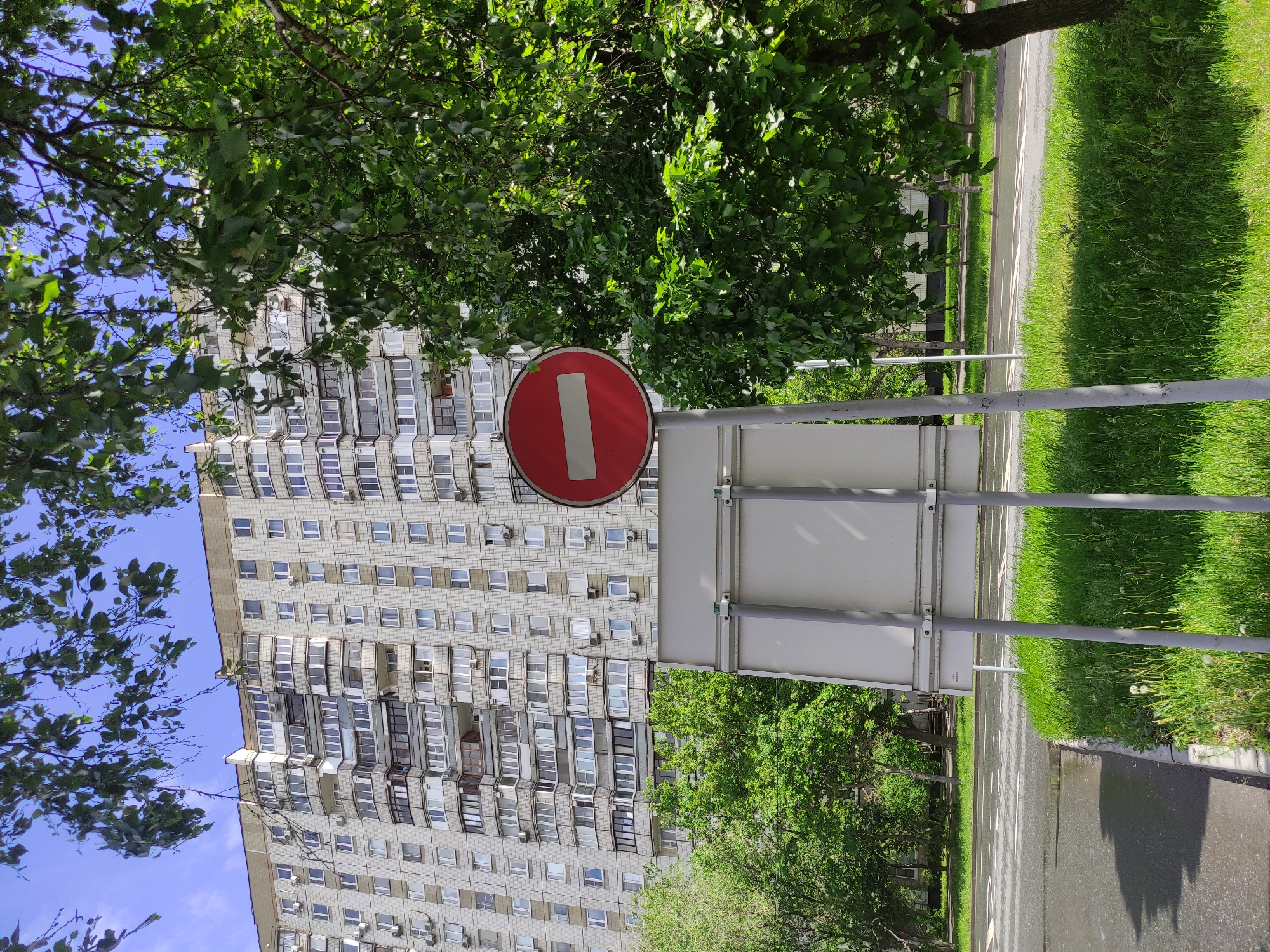The image depicts a street scene in Moscow, Russia, featuring a "No Entry" traffic sign prominently in the foreground. The sign is red with a white horizontal bar, mounted on a gray metal post. Behind the sign, a multi-story residential building with numerous windows and balconies dominates the background. Green trees surround the building and line the street, partially obscuring the view of the building. A paved road runs alongside a strip of grass, with visible road markings. The sky is partly visible through the branches of the trees, with some blue patches. The overall impression is of a typical urban landscape in Moscow.