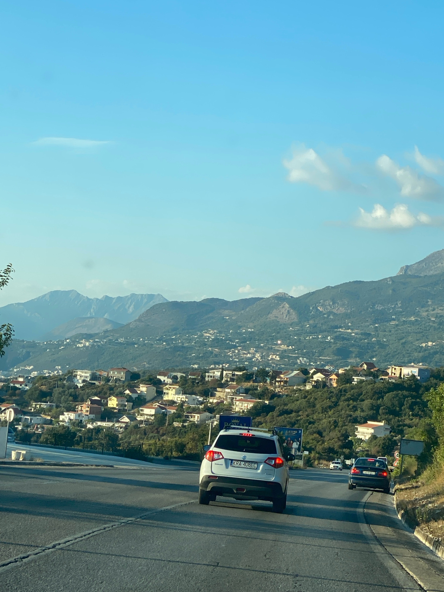 The image captures a view from a vehicle driving along a paved road in Kunje, Montenegro, on a clear day with a few scattered clouds. The road curves slightly to the right and is bordered by a grassy verge on the right side. In the foreground, the rear of a white SUV with license plate "KRA 408EC" is visible. Further ahead, a dark-colored sedan is also traveling on the road.
Beyond the road, a hillside is densely populated with numerous houses, many featuring red-tiled roofs, set amidst lush green vegetation. The houses are clustered together, creating a suburban or village-like atmosphere. In the background, majestic mountains rise, their peaks rugged and imposing, partially shrouded in a light haze. The sky is a vibrant blue. The overall scene depicts a scenic drive through a coastal area with a mix of natural beauty and human settlement.