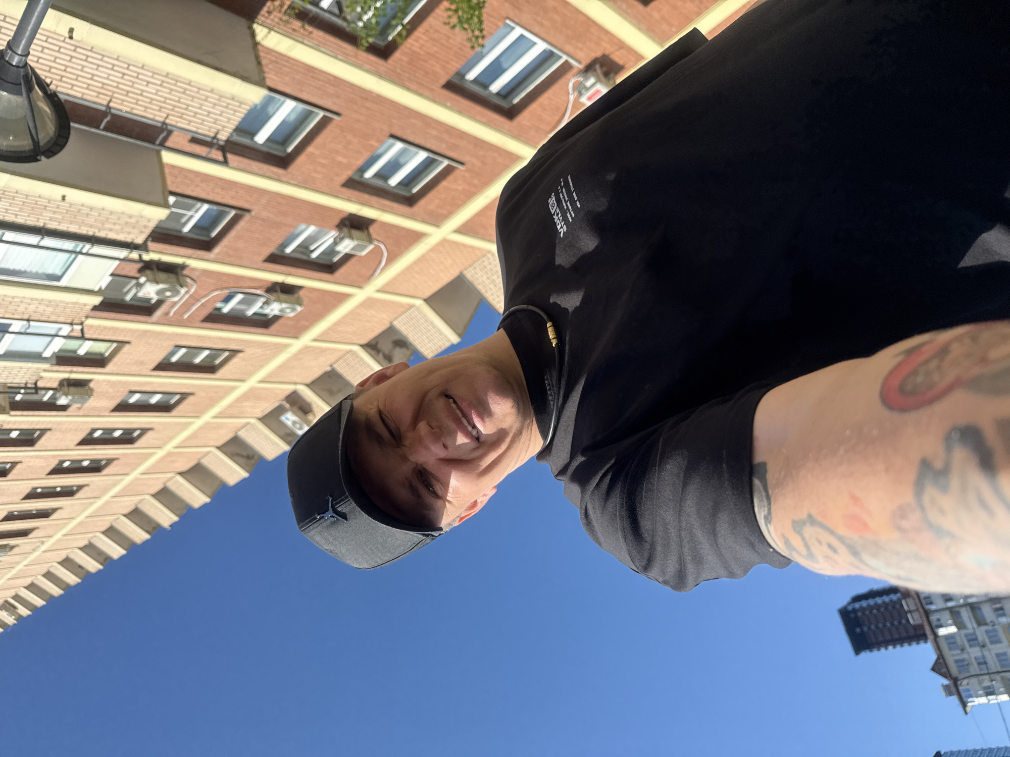 The media file shows a low-angle selfie of a man wearing a black baseball cap and a black t-shirt. He is smiling and appears to be in good spirits. His left arm, which is visible on the right side of the frame, has tattoos. The background consists of a clear blue sky and the facades of brick buildings. The buildings are multi-story with visible windows and architectural details. The lighting suggests it is daytime. The image is oriented with the top of the frame pointing downwards. The location context is Vladivostok, Russia. There is text visible on the man's t-shirt, appearing to be a logo or design.