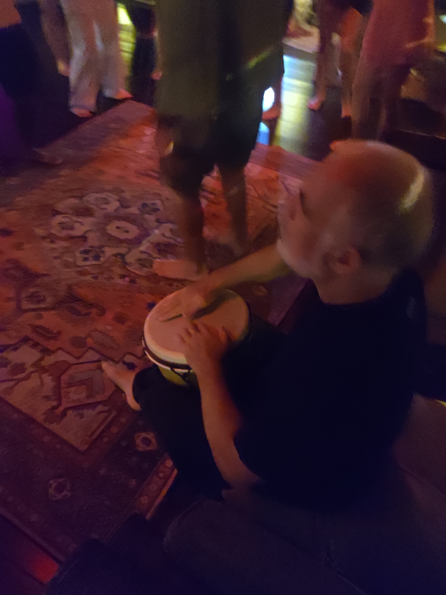 A man with a shaved head and wearing a black shirt is seated and playing a djembe drum. He is sitting on a gray cushioned surface. His bare feet are visible. A patterned rug covers the floor. Other people are standing and milling around in the background. Their clothing is varied, including pants and shorts. The location is possibly indoors, based on the lighting and rug. There is no visual information to identify Lokaserena, Indonesia.