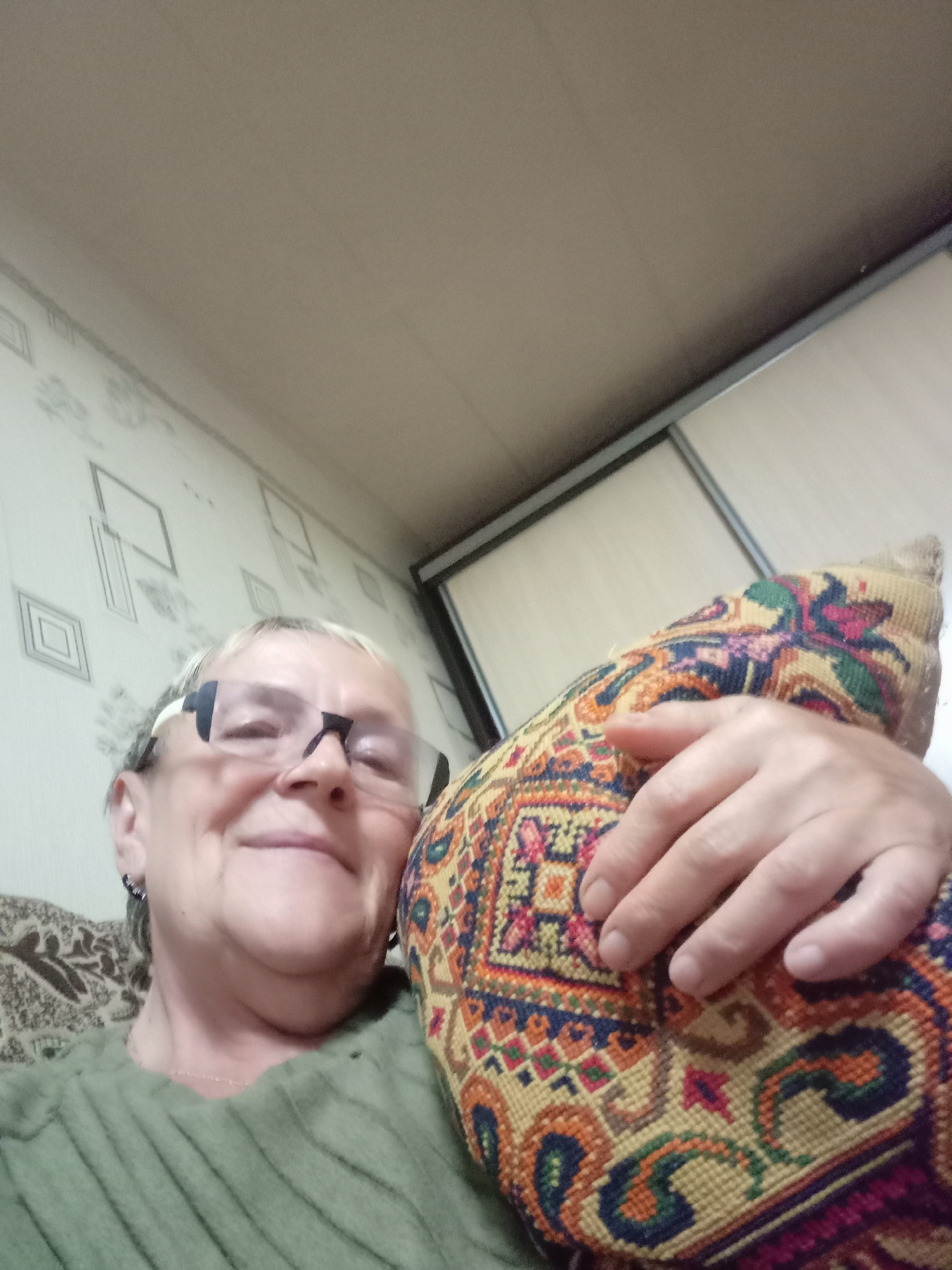 The image is a close-up selfie taken indoors of an older woman with short gray hair, wearing glasses and a green ribbed sweater. She is holding a decorative pillow with a colorful, embroidered pattern. The background appears to be a room with patterned wallpaper and a sliding wardrobe door. The lighting is warm and ambient. The scene is likely a residential interior.