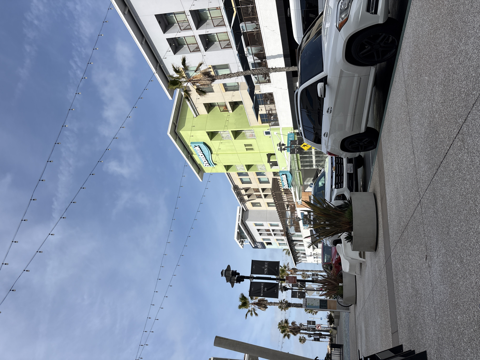 This image presents a rotated, upward-angled view of a modern street scene in Huntington Beach, California, under a clear blue sky with scattered wispy clouds. The time of day is bright daylight.
In the foreground, a light gray, speckled sidewalk stretches alongside a street where vehicles are parked. A white SUV, appearing to be a Mercedes-Benz GL-Class with black wheels, is prominent, and behind it, a California license plate reading "9YO9021" is visible on another car. Concrete planters containing spiky, ornamental plants and a white outdoor chair are placed along the sidewalk.
Overhead, multiple parallel strings of lights extend across the expansive blue sky. Palm trees, common in coastal California, punctuate the scene, standing tall between the buildings.
The architecture consists of multi-story, contemporary buildings. A distinctive light green building with a prominent blue "Shorebreak" logo suggests a hotel or resort, a detail reinforced by a smaller "hotel" sign below it. Other buildings feature gray, white, and beige facades with numerous windows and balconies. Further down the street, black banners hanging from lampposts display "WELCOME TO" and "SHOREBREAK". A yellow diamond-shaped sign depicting a person on crutches is also visible, indicating a pedestrian area or accessibility notice.
The scene captures a vibrant and modern urban environment, typical of a coastal resort town. There are no people visible, but the setup suggests a well-maintained commercial and pedestrian zone.