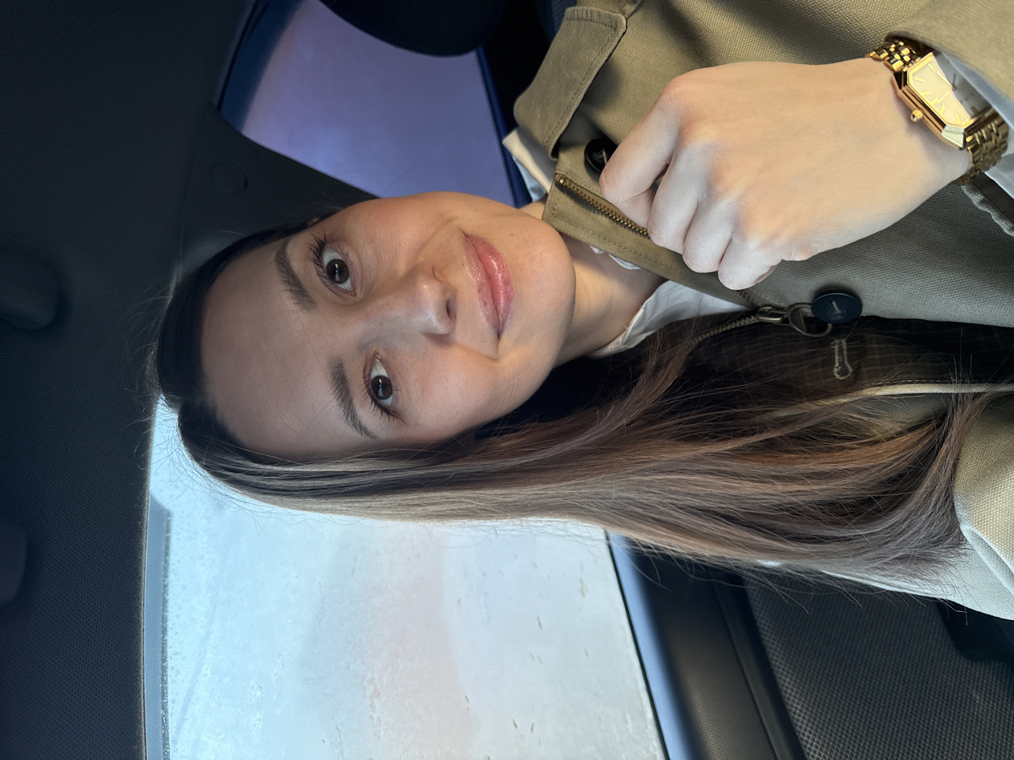 A woman with long, light brown hair poses for a selfie indoors, likely inside a vehicle, given the car window visible on the left. She wears a light green jacket with a visible zipper and a gold watch on her left wrist. The image was taken on a day with rain. The location is unknown, and there is no information to suggest this image was taken in Chaikovsky, Russia.