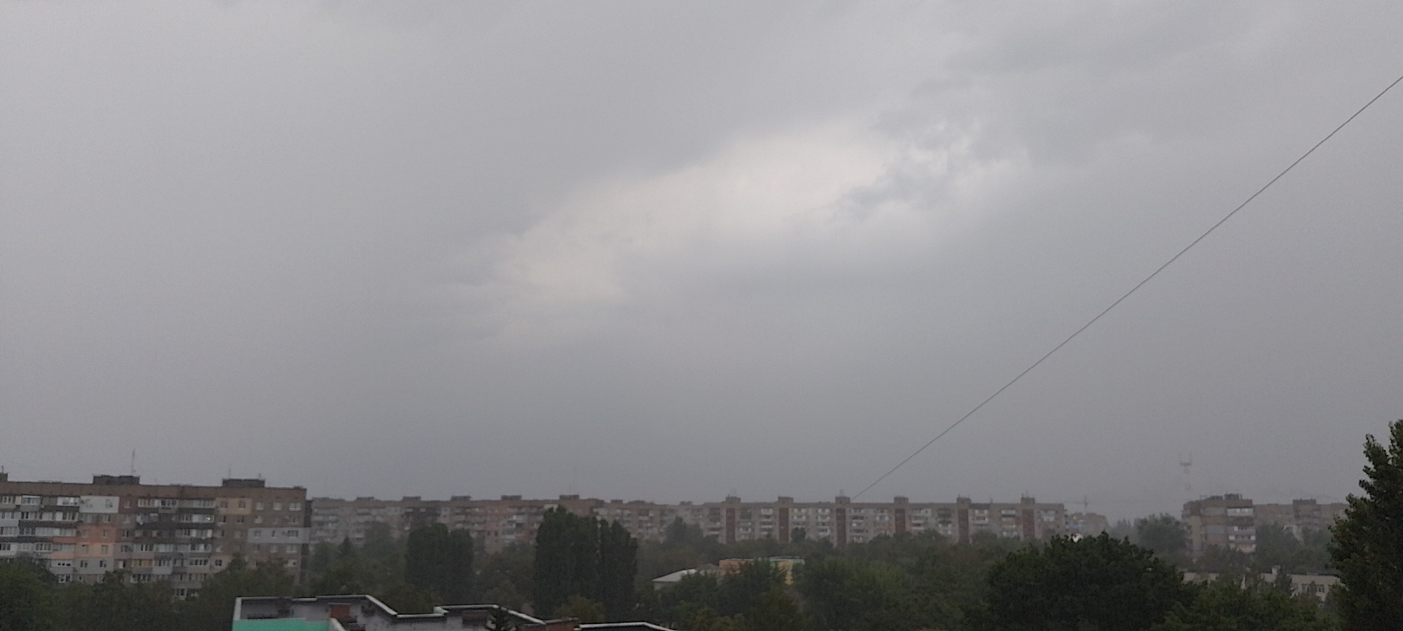 The image shows a cityscape with apartment buildings under a cloudy, overcast sky. The buildings are multi-story and appear to be typical Soviet-era residential blocks. Trees are visible in the foreground and between the buildings. A power line stretches diagonally across the sky. The overall scene suggests a slightly melancholic or subdued atmosphere, possibly due to the weather. There are no people or specific actions depicted. The perspective is from a high vantage point, looking over the city. It is not possible to determine the exact location within Cherkasy, Ukraine, from this image alone.