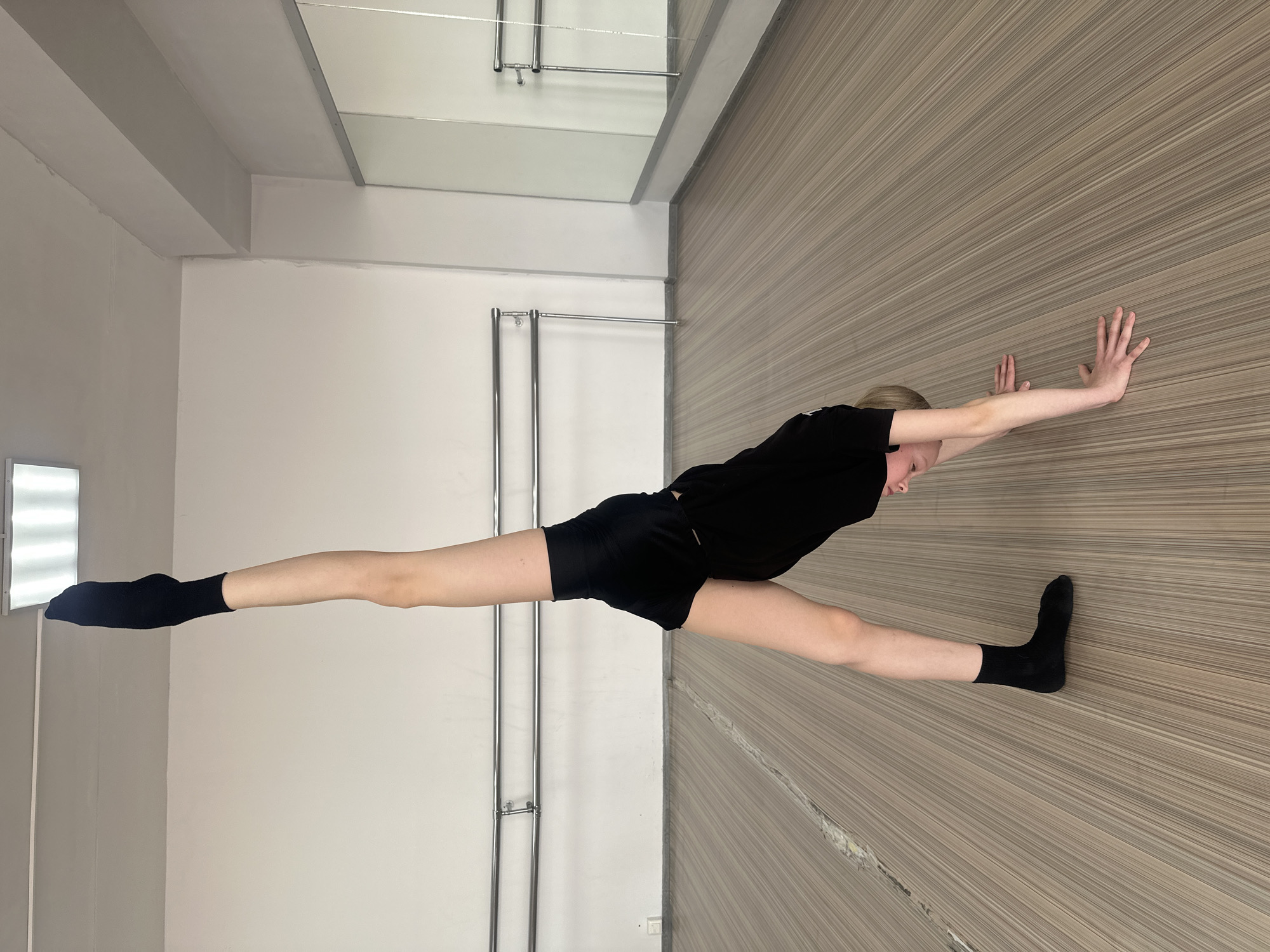 An individual is depicted performing an acrobatic or dance pose within a studio environment in Votkinsk, Russia. The subject, a young person with light-colored hair, is positioned with their torso bent forward, hands flat on the striped wood-look floor, and one leg (the left) extended vertically towards the ceiling. They are wearing a black short-sleeved top, black shorts, and black socks on both feet. The studio features white walls, a light-colored ceiling with a rectangular fluorescent light fixture, and two parallel silver metal barres mounted on the left wall. A wall of mirrors with silver framing is visible on the right, reflecting portions of the room and the barres. The floor is composed of light brown planks with fine parallel lines.