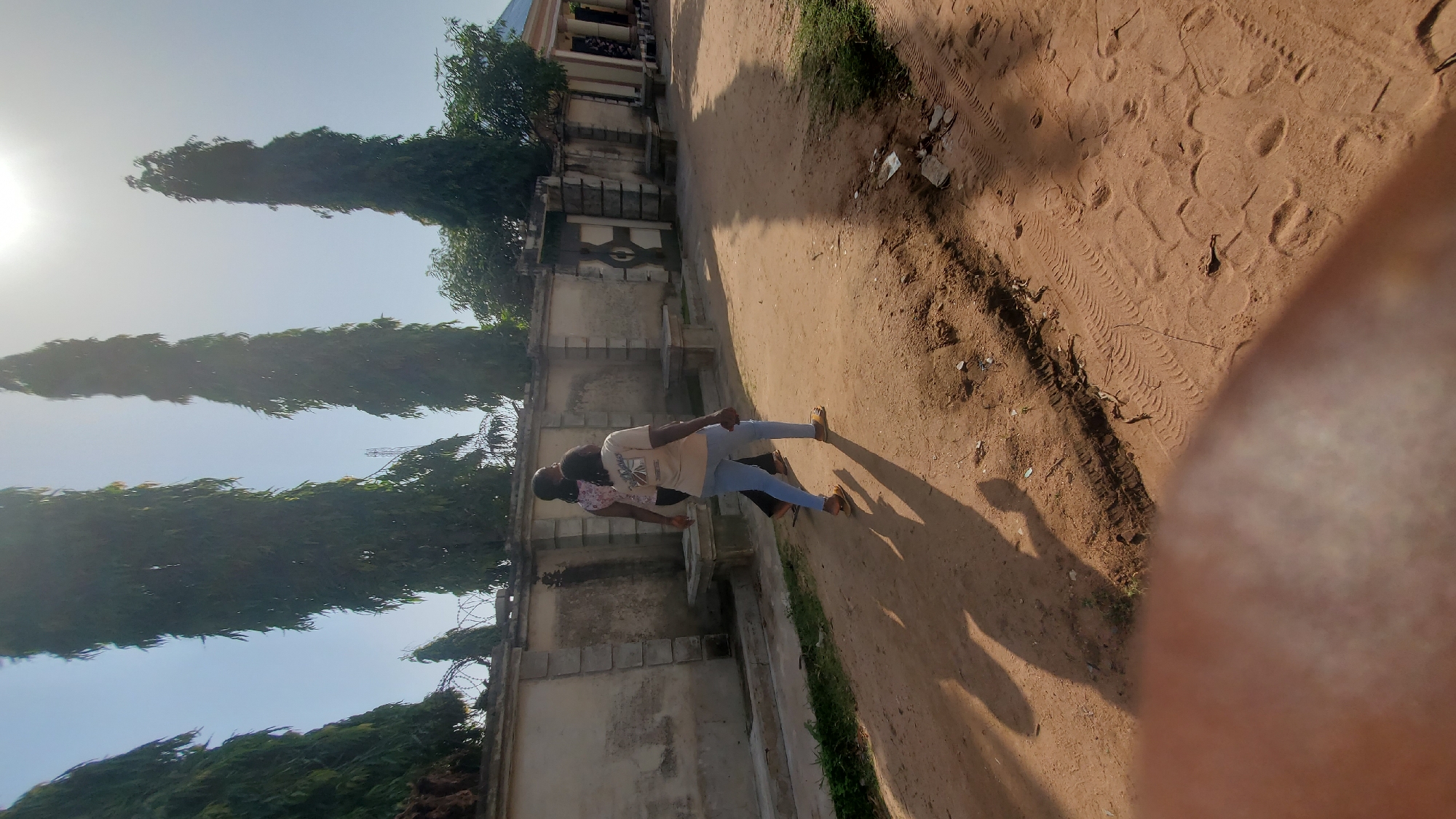 The image captures a sunny outdoor scene, likely in Jalingo, Nigeria, featuring two individuals walking along a sandy dirt path. The path appears to be a road or walkway. On the left side of the path, there's a concrete wall with decorative elements and barbed wire atop it, suggesting a property boundary. Several tall, slender trees line the top of the wall, their branches reaching towards the bright sky, which is partly obscured by the sun's glare. The ground is covered in loose sand and has tire tracks visible, indicating vehicle passage. In the foreground, a blurry, out-of-focus area of what appears to be skin or fabric occupies the bottom left corner of the frame. The two individuals, seen from behind and walking away from the camera, are dressed casually; one is wearing a light-colored t-shirt with a graphic design and blue jeans, while the other wears a patterned shirt and dark trousers. Their shadows stretch long across the sandy ground, indicating a low sun angle. The overall environment suggests a residential or public area with substantial vegetation and a clear sky.