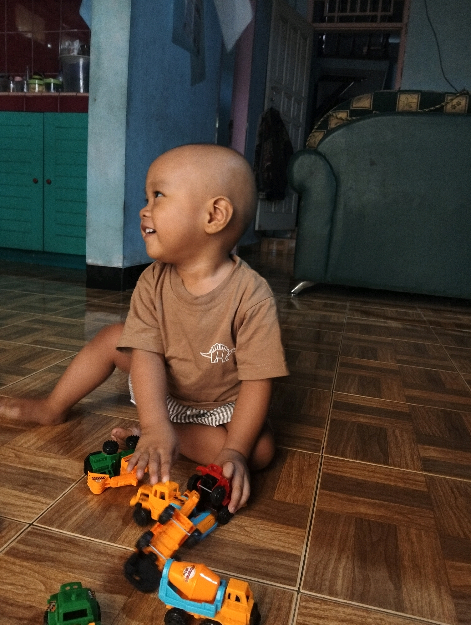 A young child with a shaved head, wearing a brown t-shirt with a dinosaur print and striped shorts, sits on a tiled floor, playing with several toy trucks. The child is facing left, with a slight smile on their face. The toys include a green truck, an orange dump truck, and a cement mixer truck. In the background, there is a dark green sofa, a wooden door, and a turquoise cabinet. The flooring appears to be wood-patterned tiles. The scene suggests a domestic setting, likely within a home in Adinuso, Indonesia.