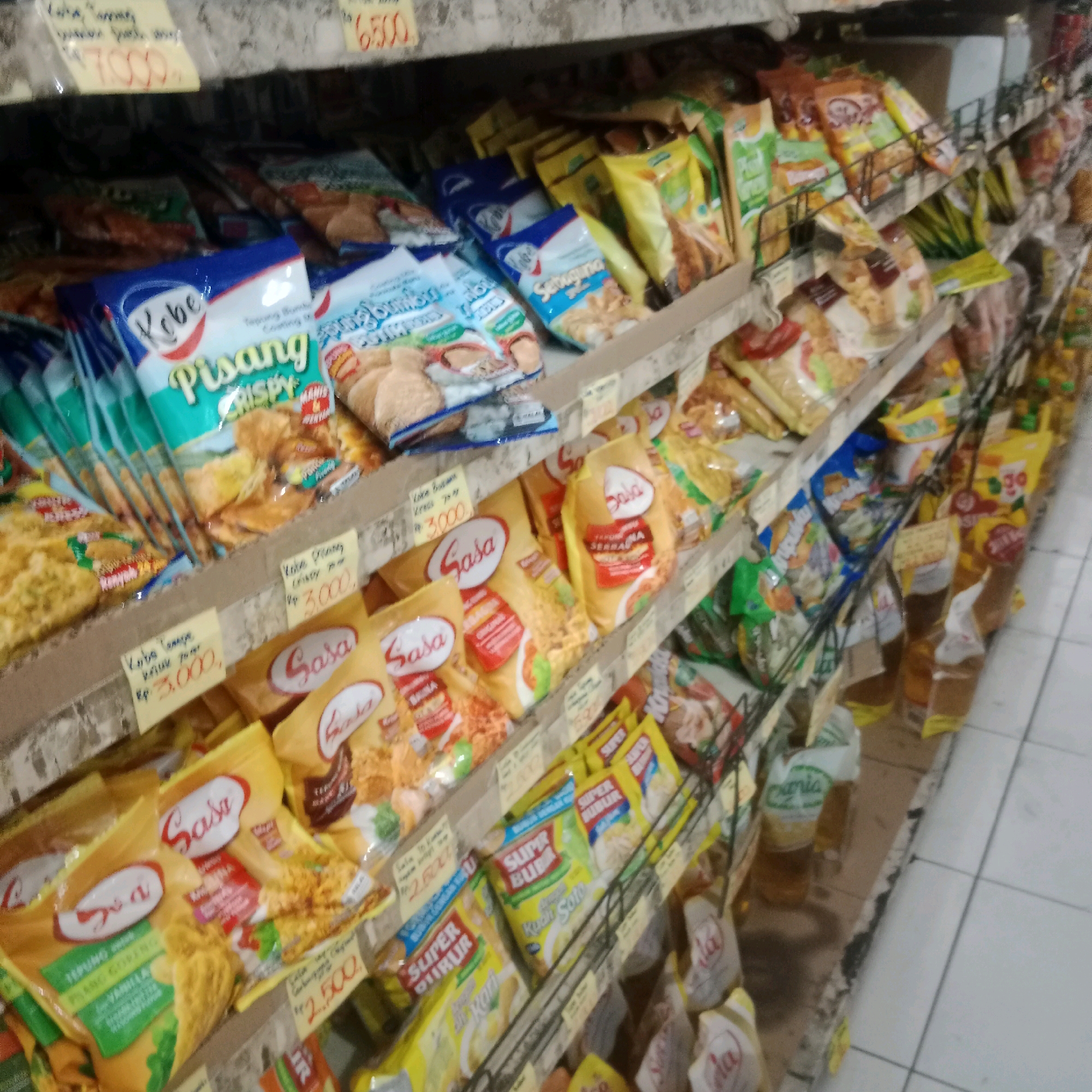 The image depicts a shelf in a small grocery store, likely a warung or minimarket, in Tegowanu Wetan, Indonesia. The shelves are filled with various packaged food items, predominantly snacks and instant noodles. In the foreground, a shelf displays multiple yellow packets of "Sasa" brand seasoning and crispy flour. To the left, there are blue and green packets labeled "Kobe" and "Pisang Crispy". Further back and to the right, there are more rows of packaged goods, including what appear to be instant noodle packs like "Super Bubu" and other unidentifiable snacks. Several yellow price tags are visible on the shelves, with handwritten prices such as "Rp 3.000," and "Rp 2.500,". The lighting is dim, and the overall scene suggests a typical retail environment. The floor is tiled with white and light grey squares, and the shelves are made of metal with wooden support structures. The image is shot from a slightly elevated angle, looking down onto the shelves. There are no people or animals visible in the image.