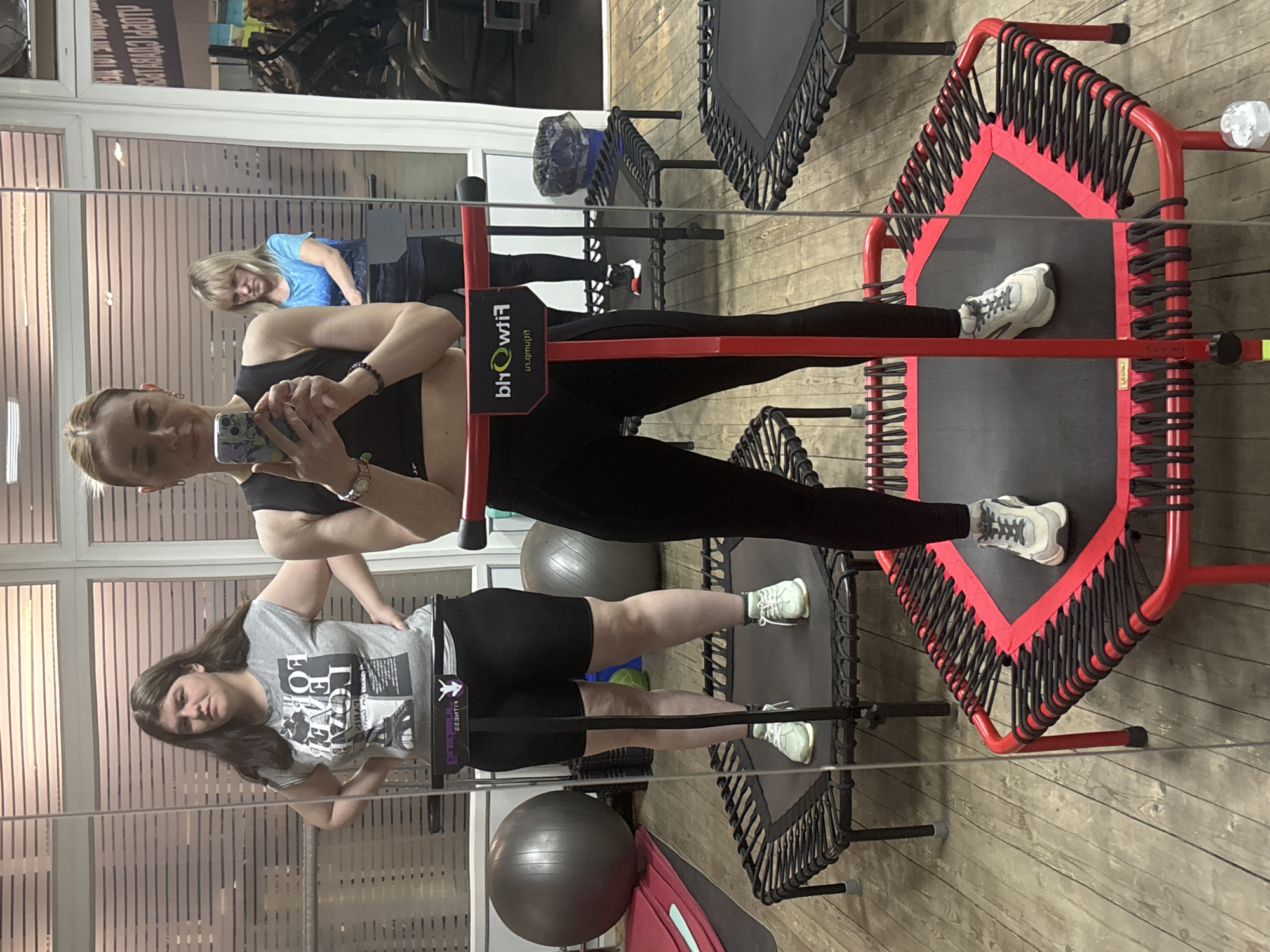 The image shows two women in a gym setting, likely in Saint Petersburg, Russia based on the Cyrillic text visible in the mirror reflection. The woman closest to the camera is taking a selfie, holding her phone up. She is wearing a black sports bra and black leggings. Behind her, reflected in the mirror, another woman with blonde hair is wearing a blue shirt. Below the first woman, another woman is reflected in the mirror, wearing a grey t-shirt and black shorts. Both women in the reflection appear to be on mini-trampolines with handlebar supports. A red and black trampoline is visible on the right, while a black trampoline is visible further in the background. Gray exercise balls are also visible. The gym has a wood-patterned floor.